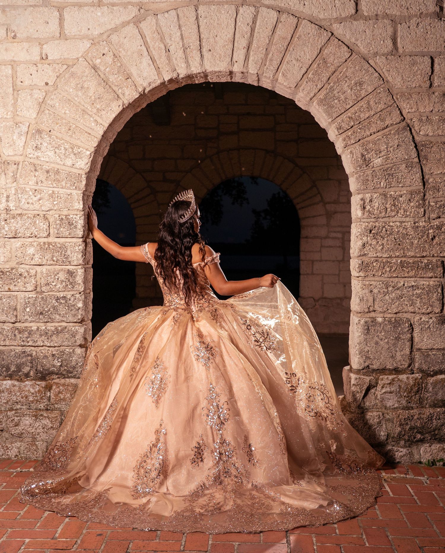Elegant ball gown with floral appliques and glittering details displayed in archway of historic stone architecture.