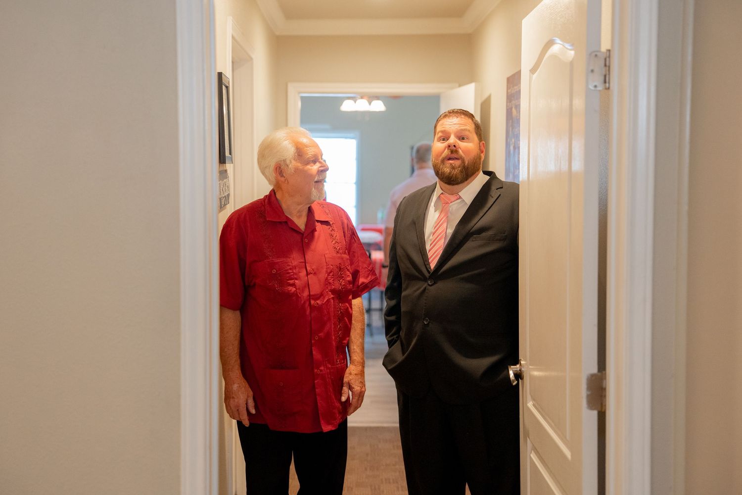 Two men standing in a hallway, one in a red shirt and one in a gray suit, chatting at a wedding.