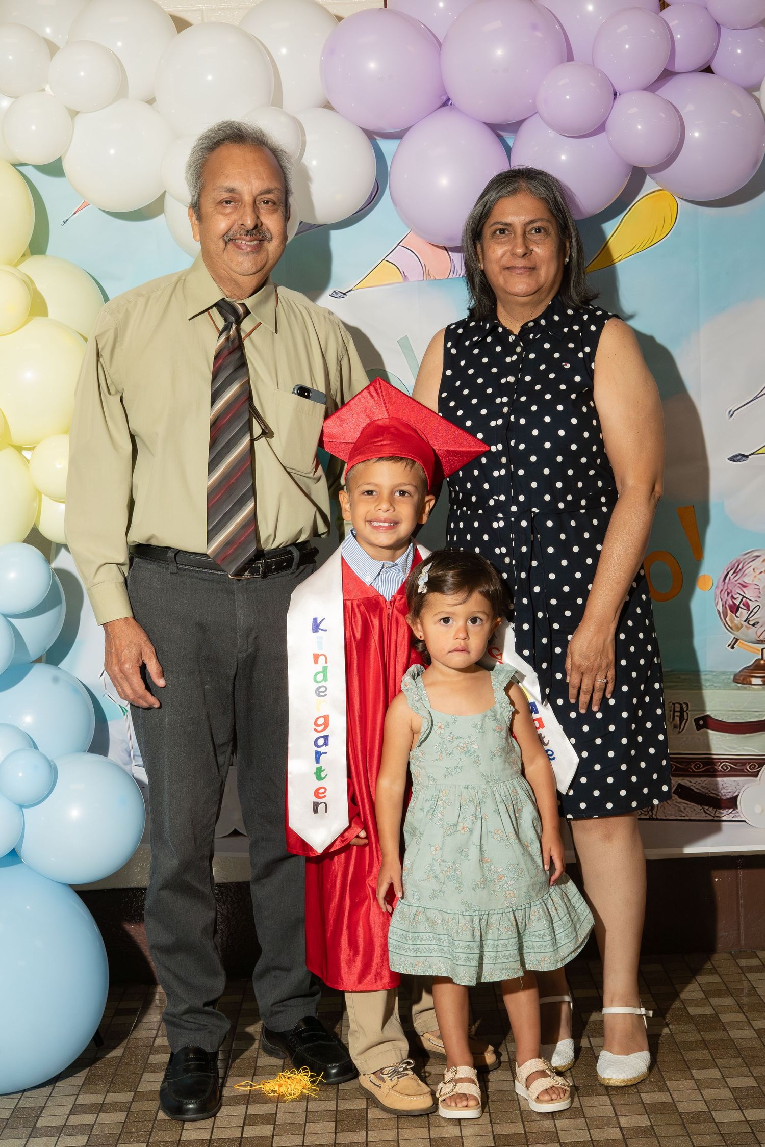 Family photo at a graduation celebration with balloons, featuring a child in red cap and gown with parents and sibling.