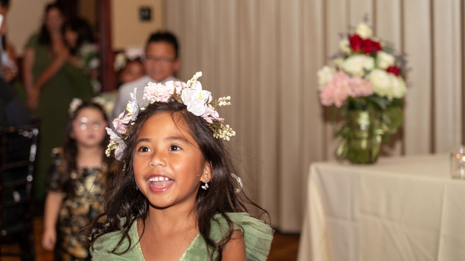 Young girl in floral crown walks at wedding ceremony in elegant venue with flowers and guests.