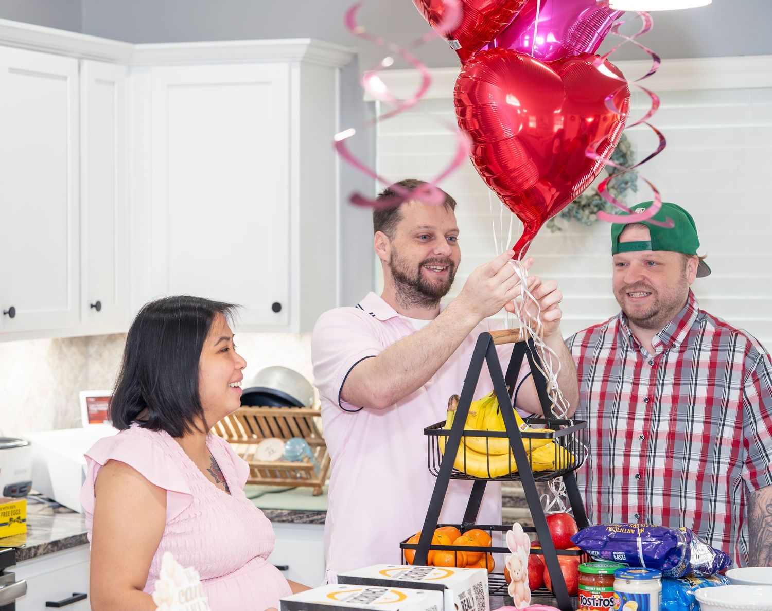 Two men and a woman celebrate in a kitchen with red heart-shaped balloons on a ladder centerpiece.