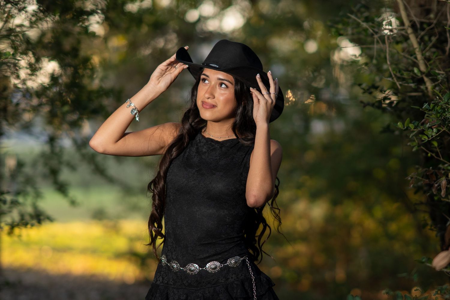 Young woman in black dress and cowboy hat posing outdoors among green trees in a stylish fashion portrait.