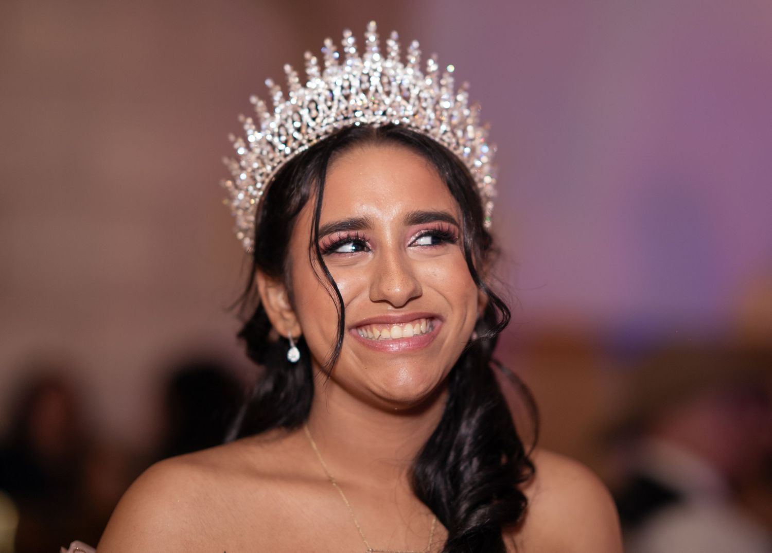 A beautiful Quinceañera smiles radiantly wearing an elaborate pearl and crystal crown at her celebration.