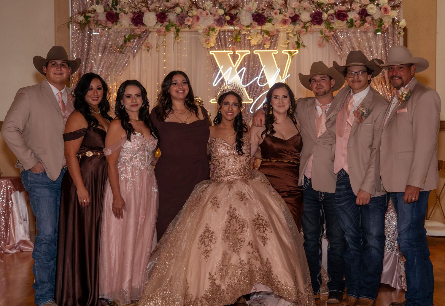 Group photo at a quinceañera celebration with guests in cowboy hats and formal gowns posing before a floral backdrop.