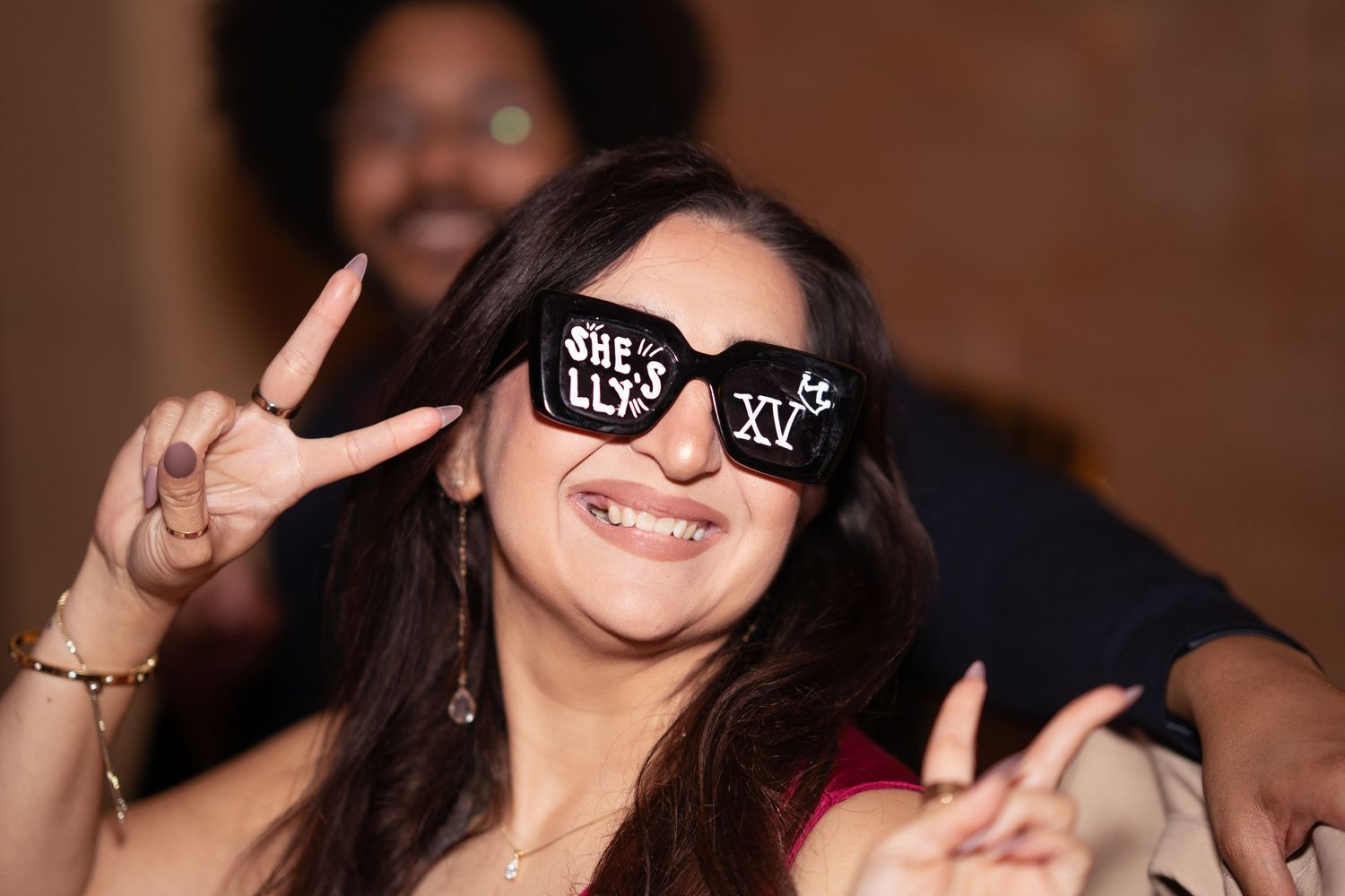 Woman smiles wearing novelty sunglasses reading She Llys XV at a quinceañera party.