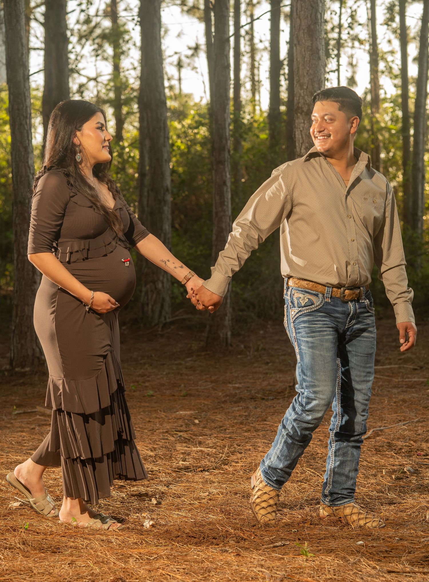 Pregnant woman in brown dress holds hands with smiling man in jeans walking through pine forest during maternity photoshoot.