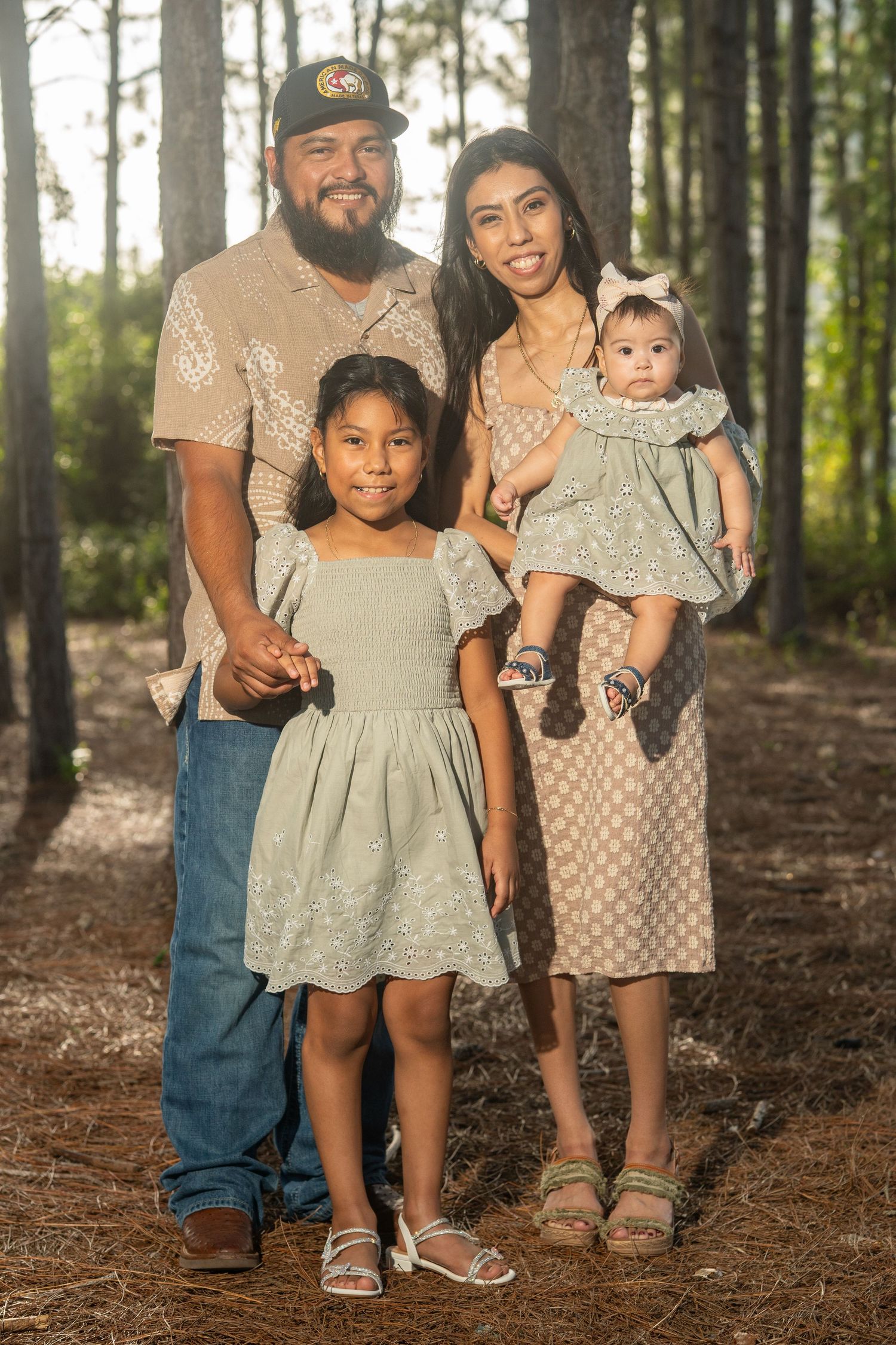 Family of four poses in pine forest, parents with two young daughters wearing coordinated sage green and beige outfits.