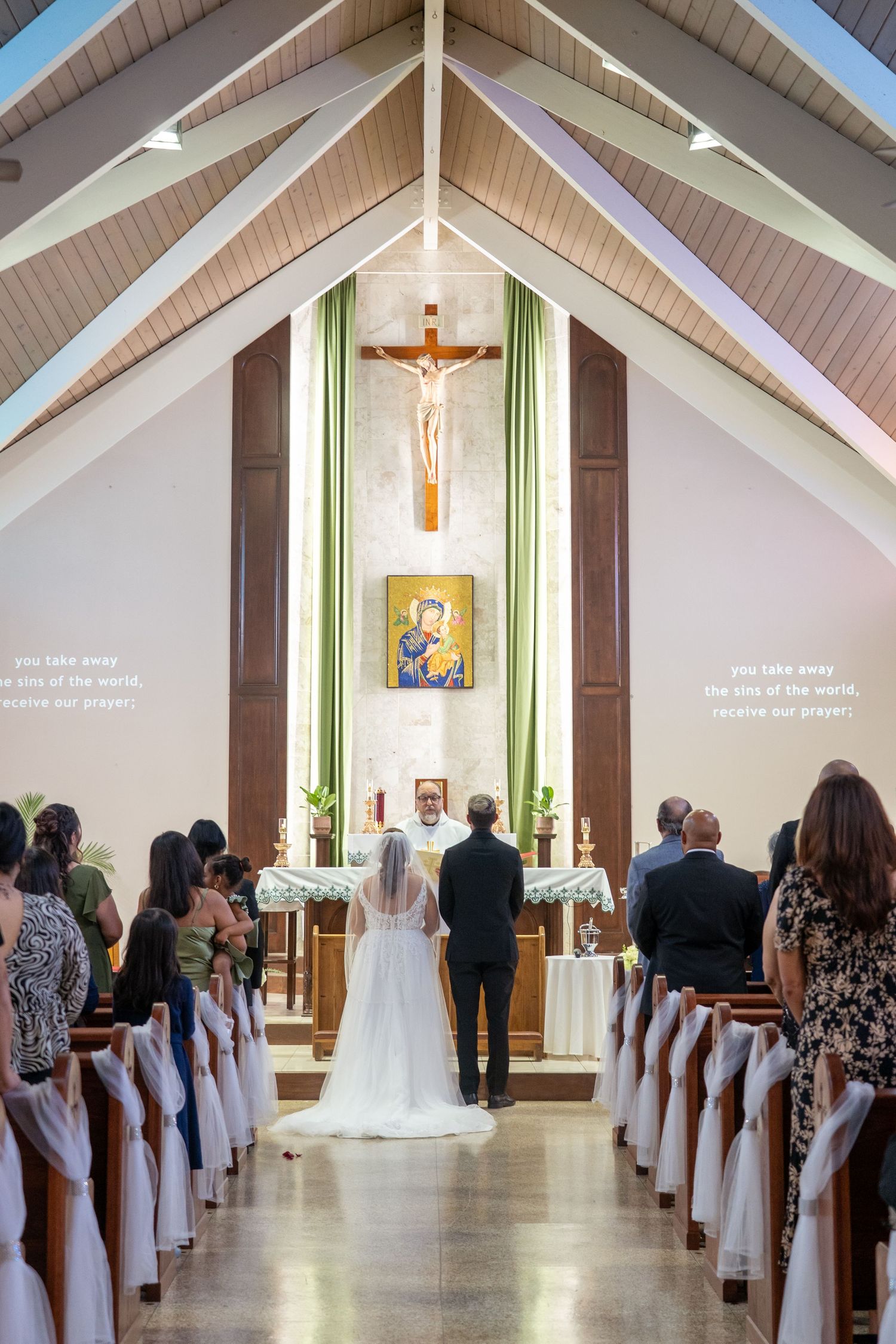 Bride and groom stand at church altar during Catholic wedding ceremony with crucifix and guests seated in pews.
