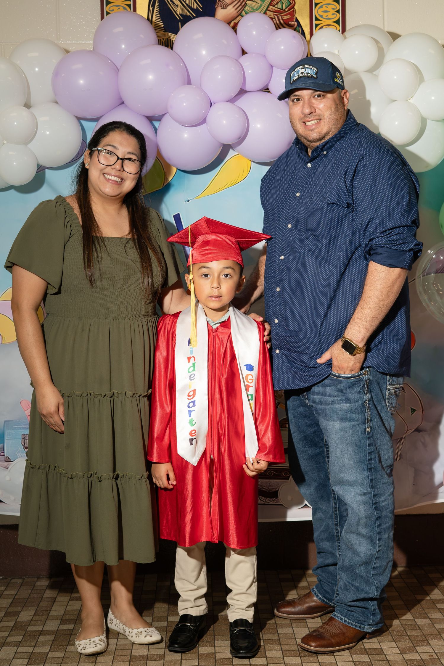 Young boy in red graduation cap and gown poses with proud parents in front of balloon backdrop.