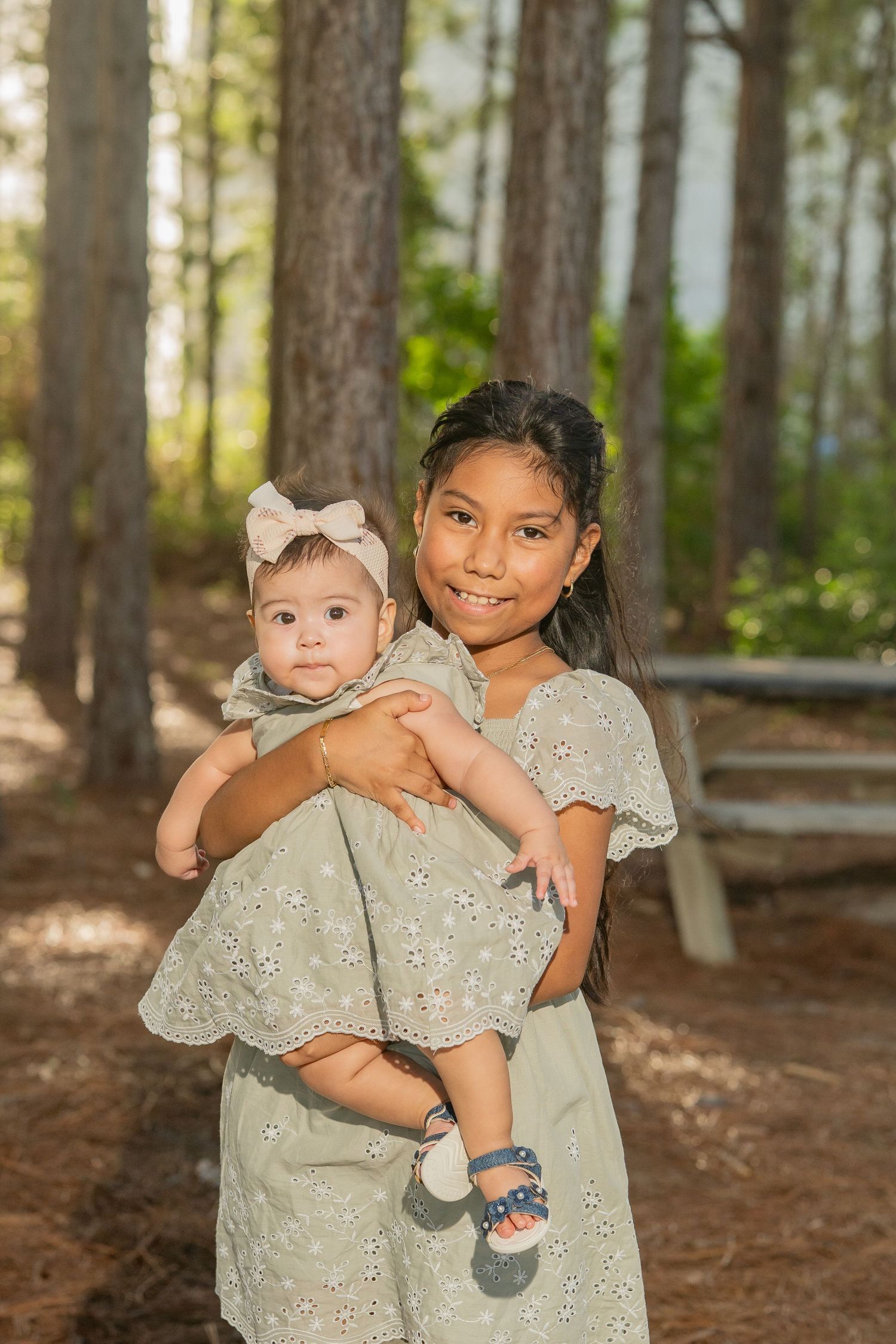 Young girl in sage green dress holds smiling baby with bow headband outdoors among tall pine trees.