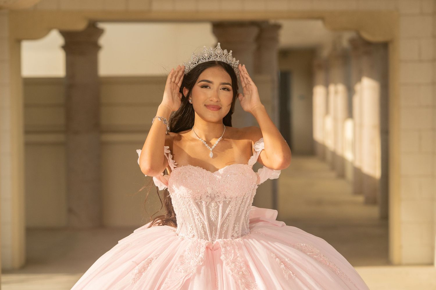 Young woman in pink ball gown and tiara adjusts crown, posing elegantly in a columned outdoor setting.