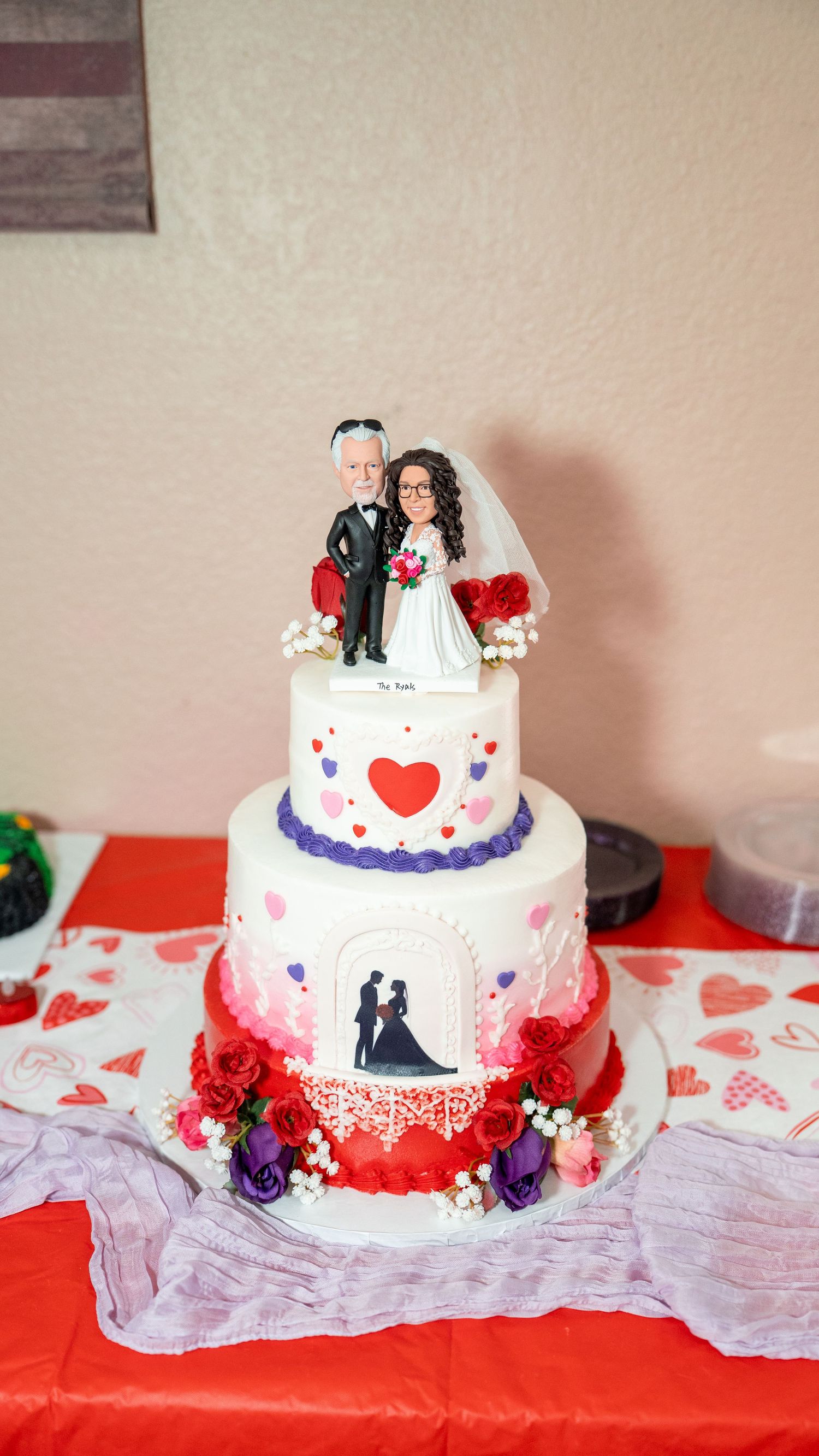 Three-tier white wedding cake with heart decorations, custom bride and groom topper, red roses and purple flowers.