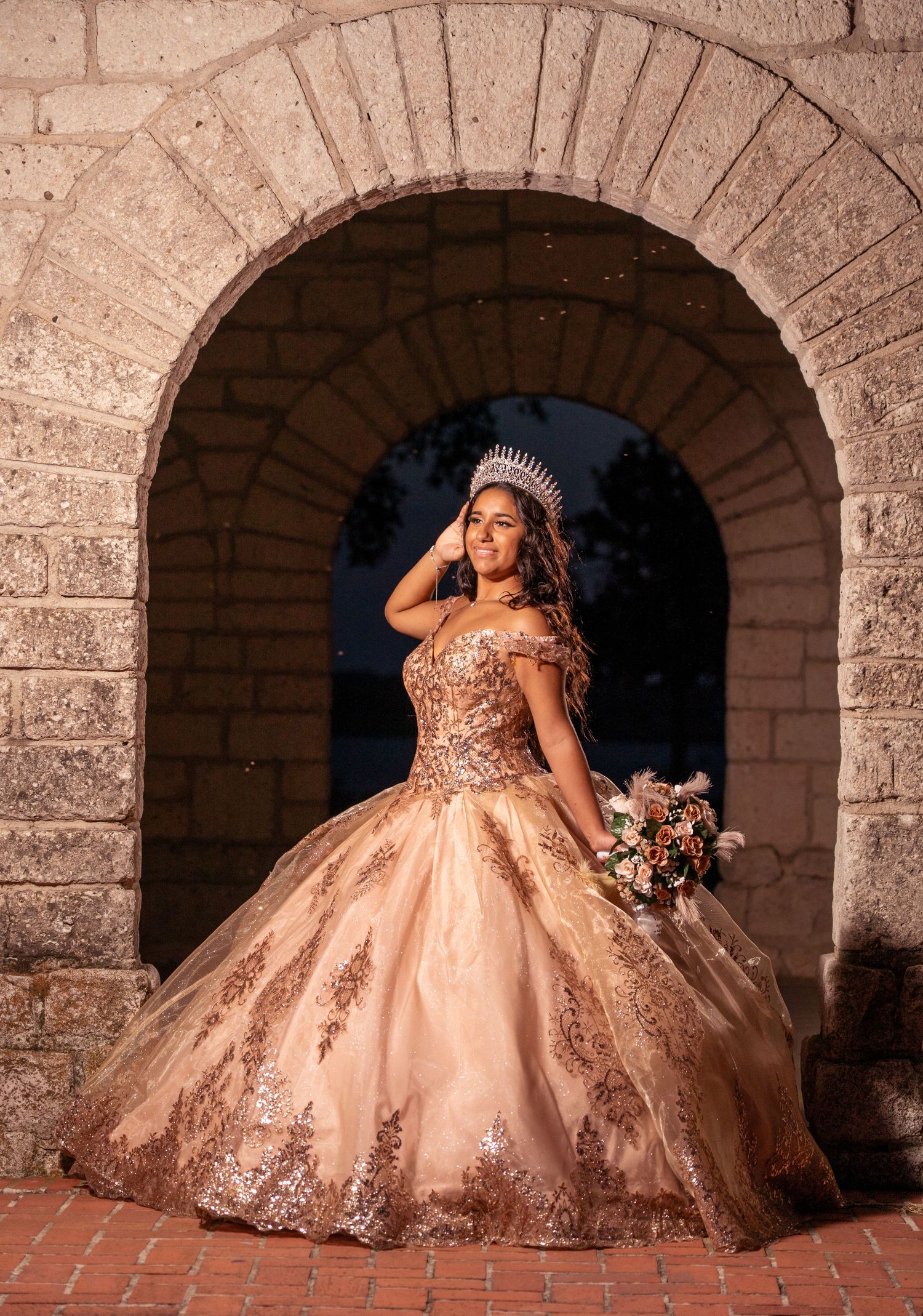 A glamorous ballgown with beaded bodice and full skirt photographed under a rustic stone archway.