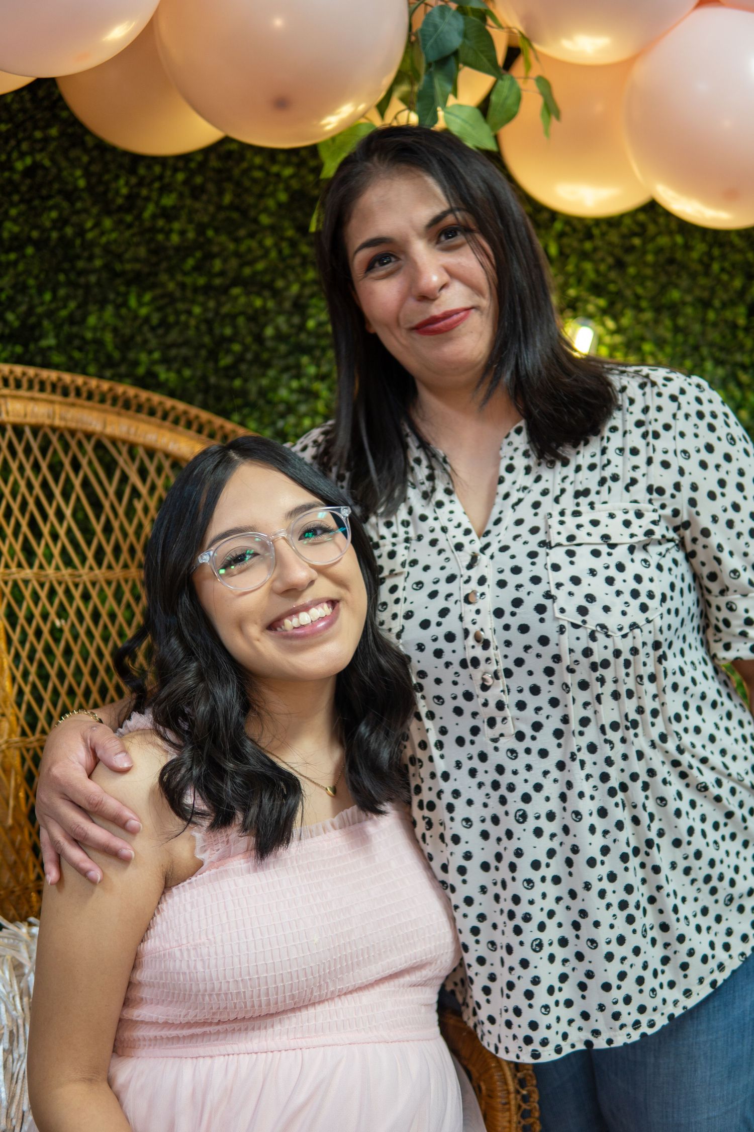 Two people smile warmly while posing together against a green wall backdrop with decorative balloons and string lights.