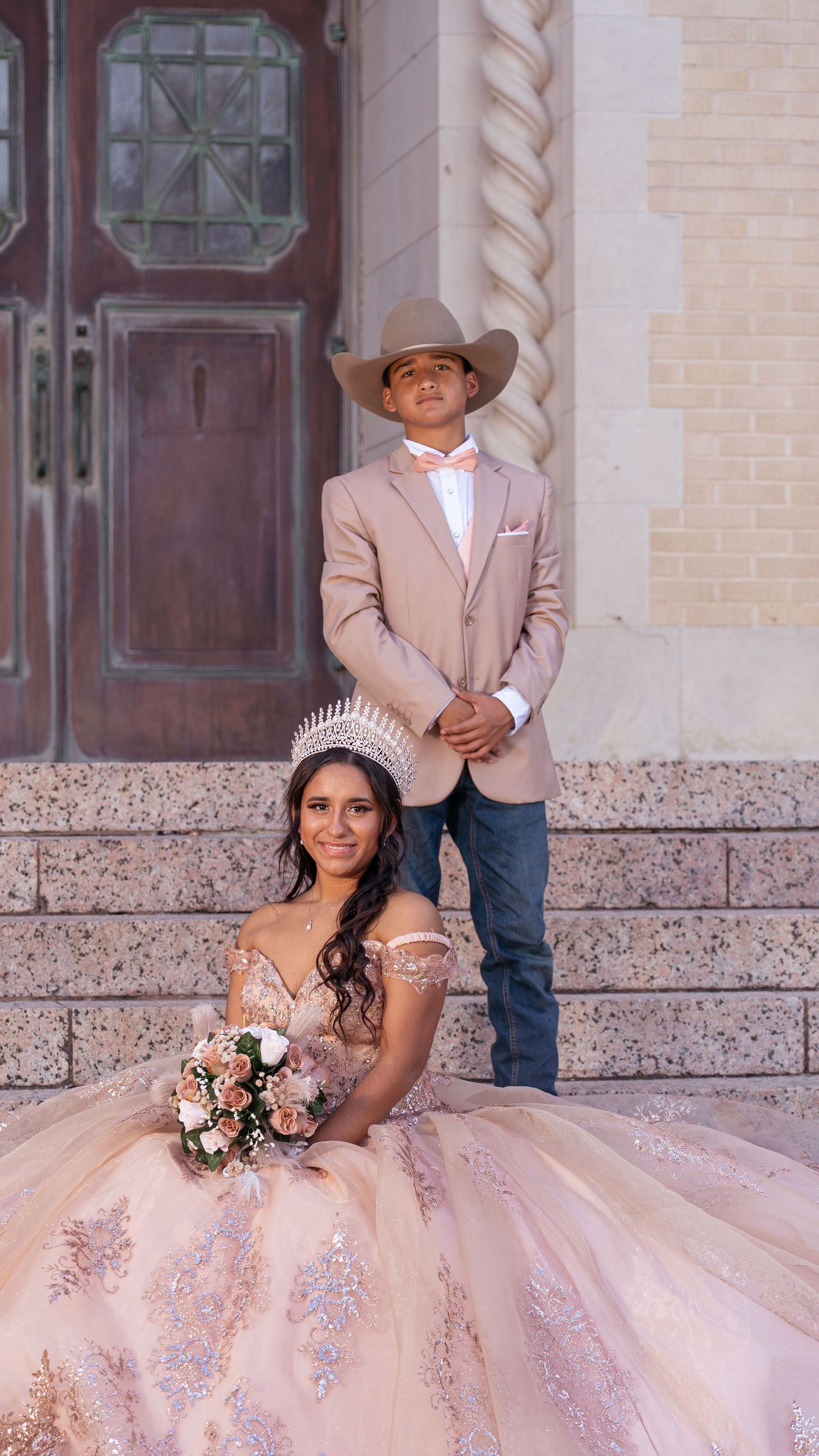 A quinceañera photo shoot featuring a pink ballgown and cowboy-themed attire against a beige stone building exterior.