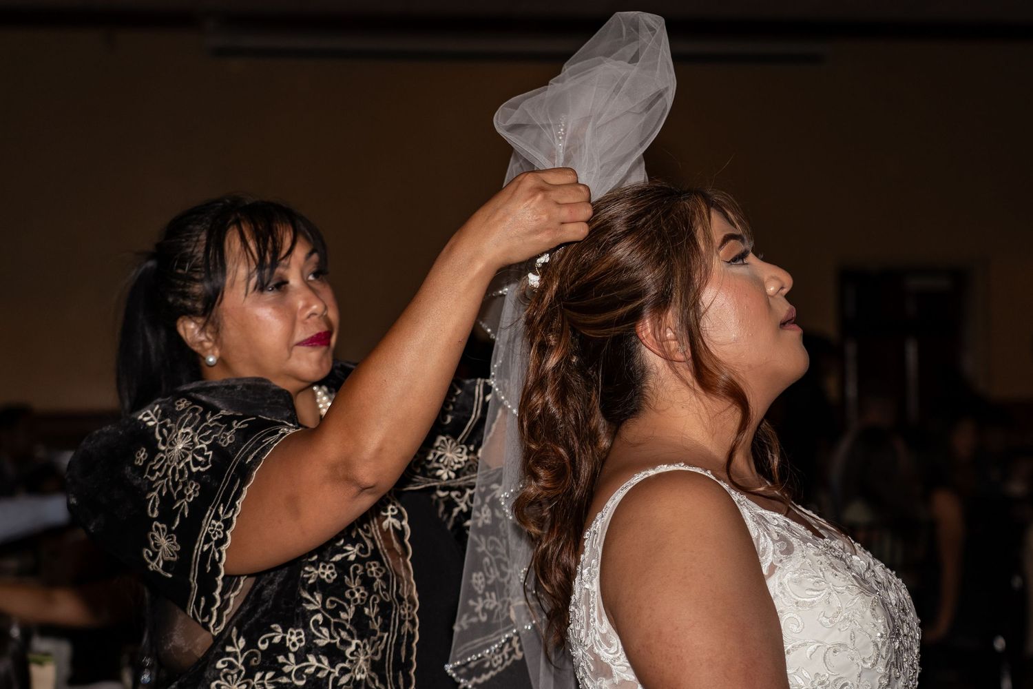 A woman carefully places a wedding veil on a bride in a white gown during a wedding reception.