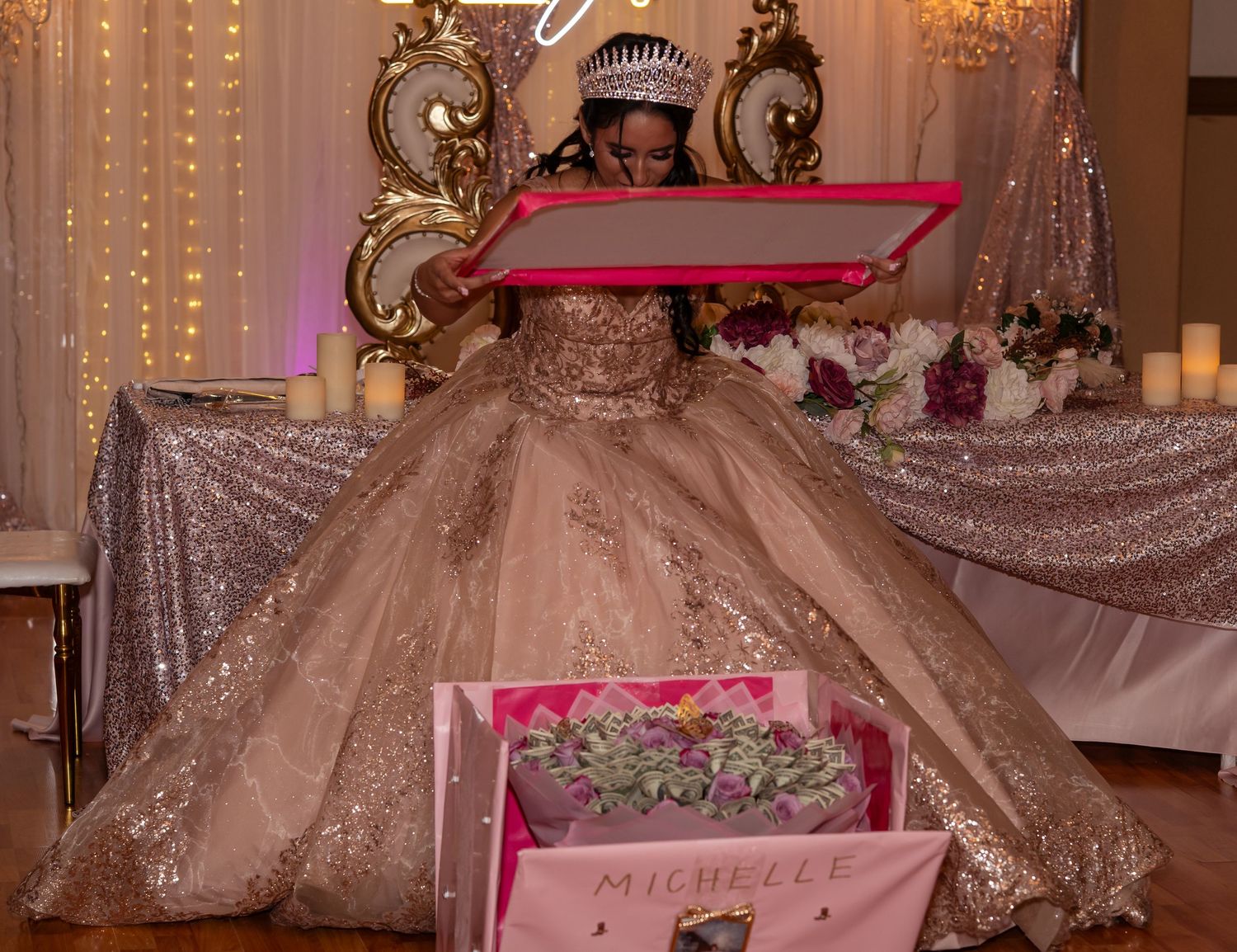 Young woman in a rose gold ball gown opens a pink gift box at her quinceañera celebration with her name Michelle.