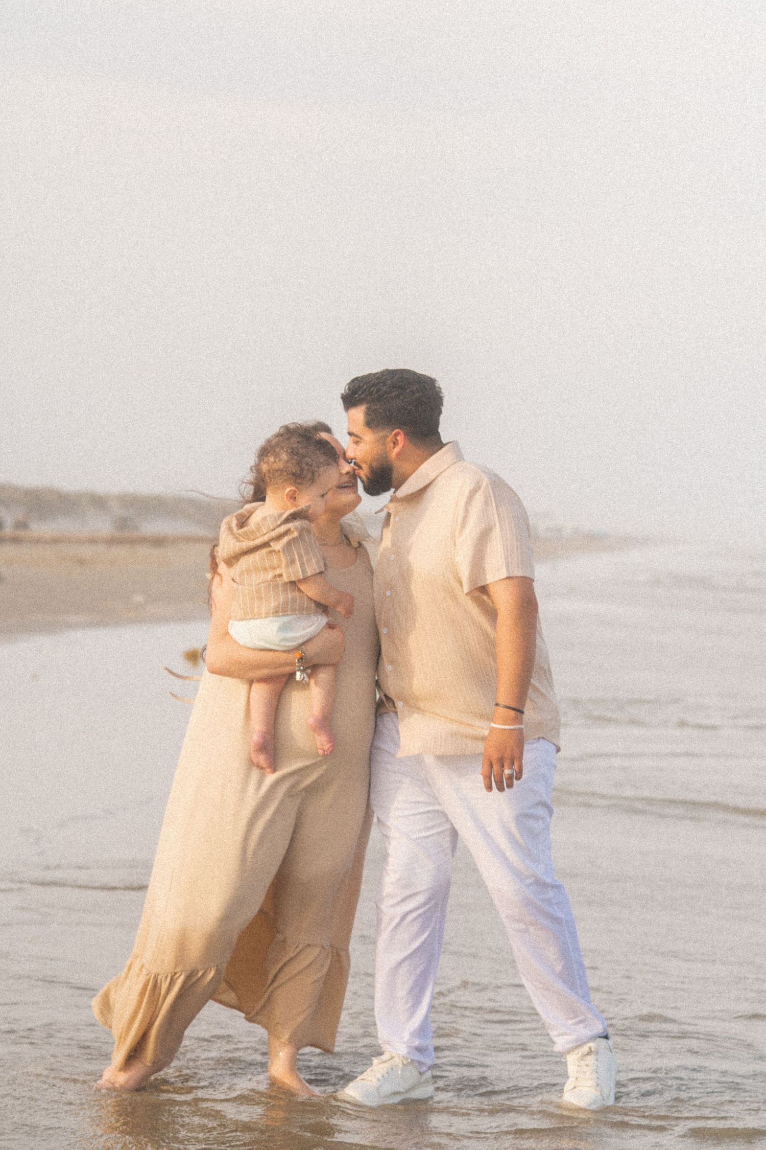 A couple in beige outfits walks barefoot on a beach, sharing a tender moment while holding their baby.