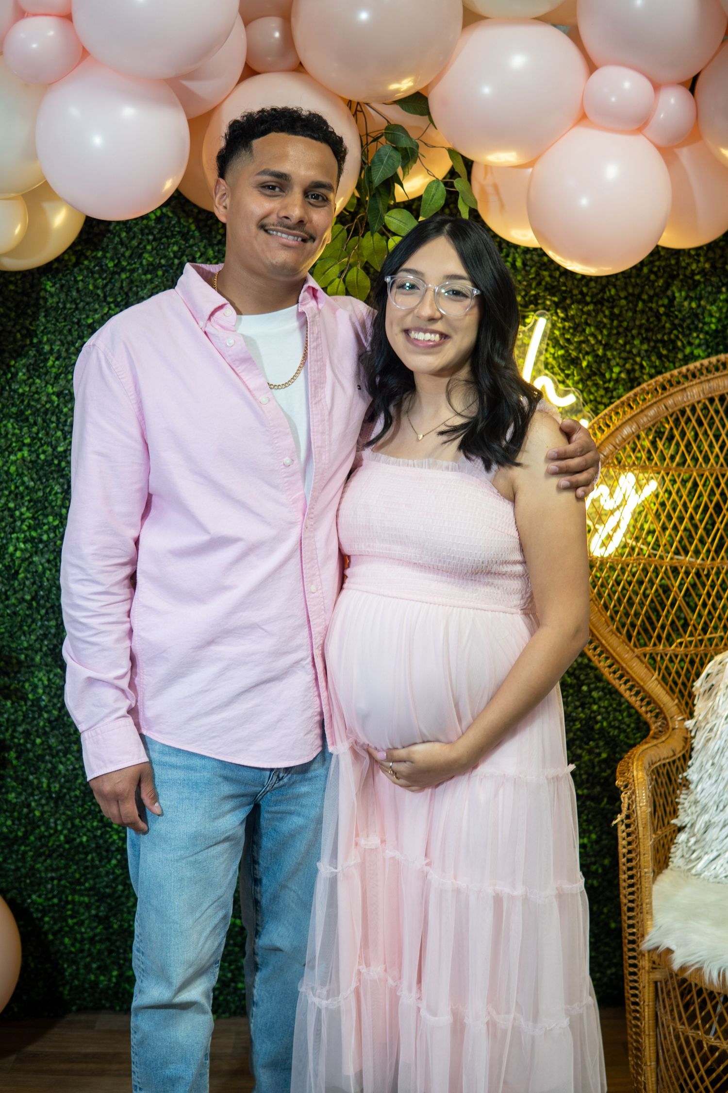 A couple poses at a baby shower celebration with pink and gold balloons and a decorative green wall backdrop.