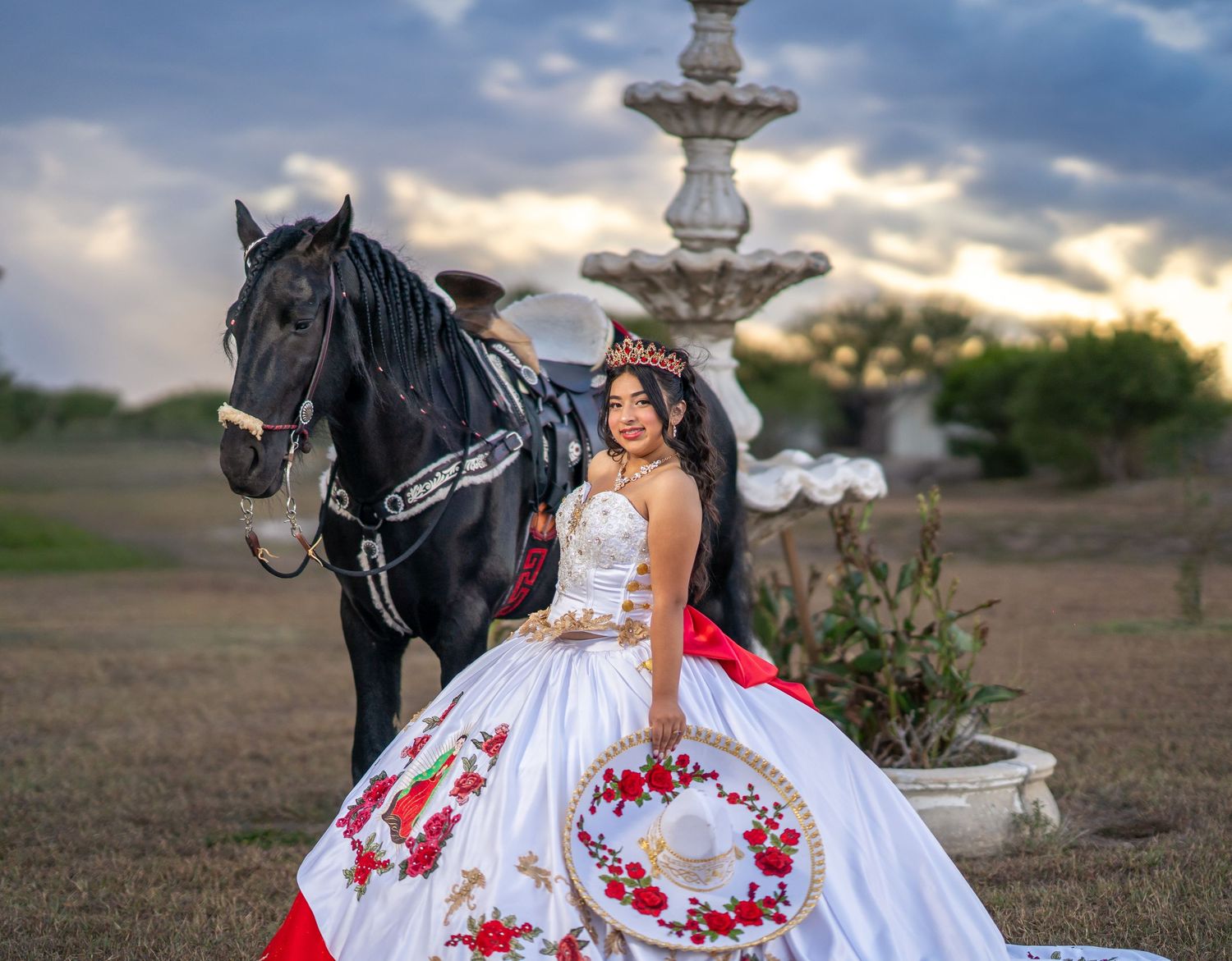 A white quinceanera dress with red floral embroidery is showcased in an outdoor photoshoot with a black horse.
