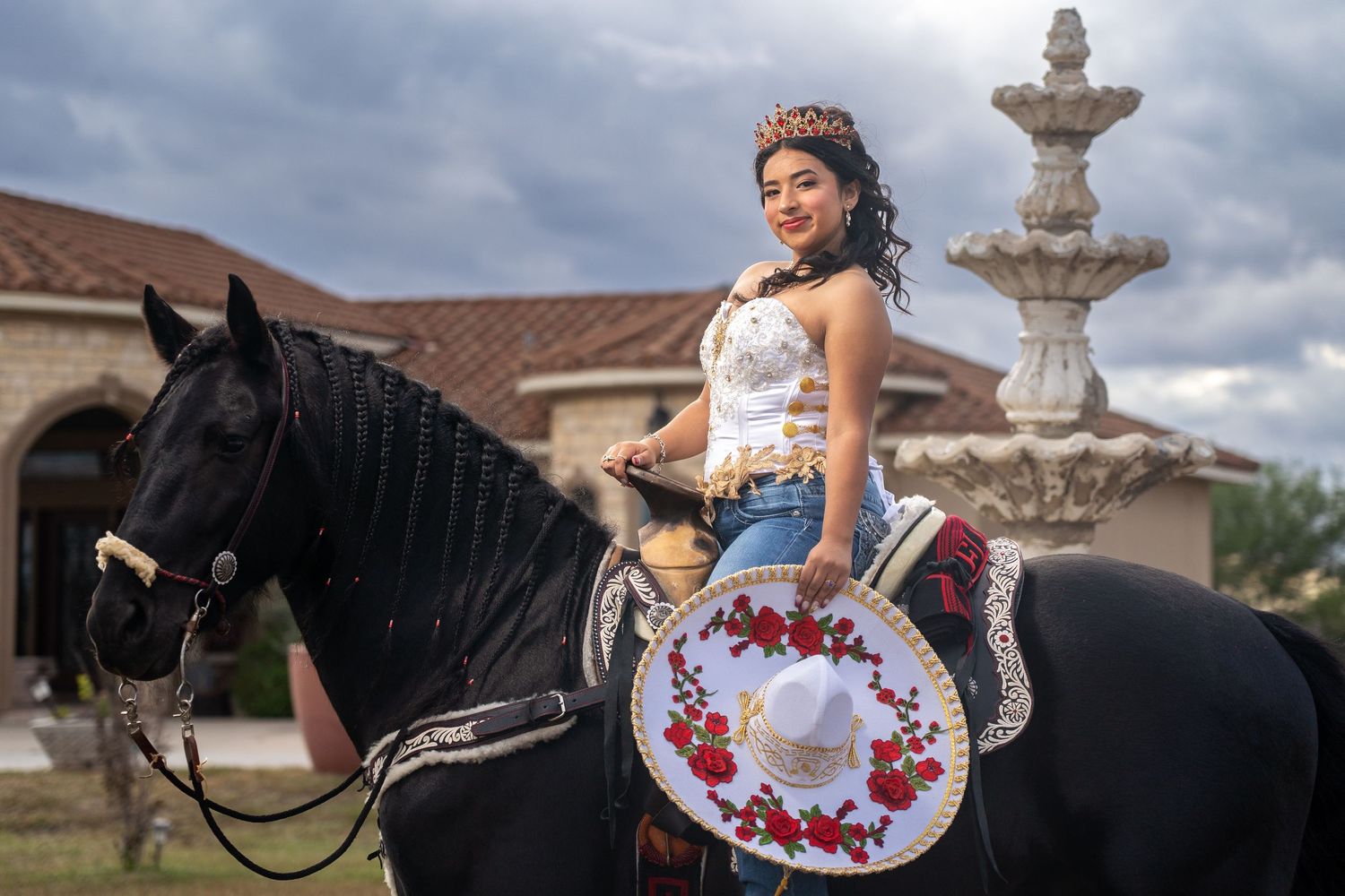 A rider in an embroidered dress sits atop a black horse near a fountain and Spanish-style architecture.