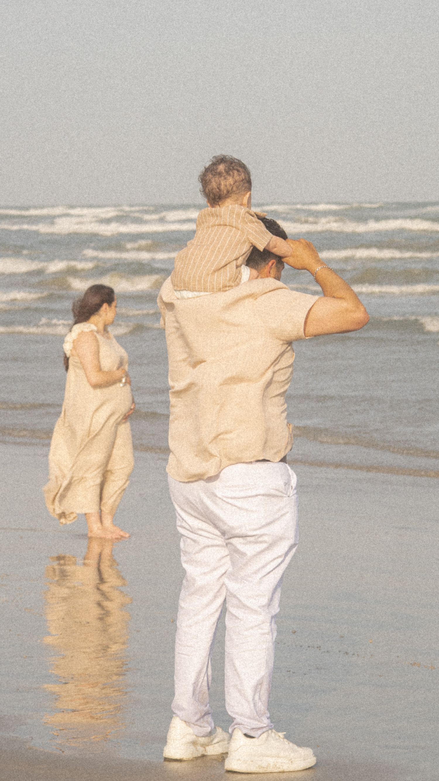 Family at the beach, father carrying child on shoulders while mother walks near ocean waves.