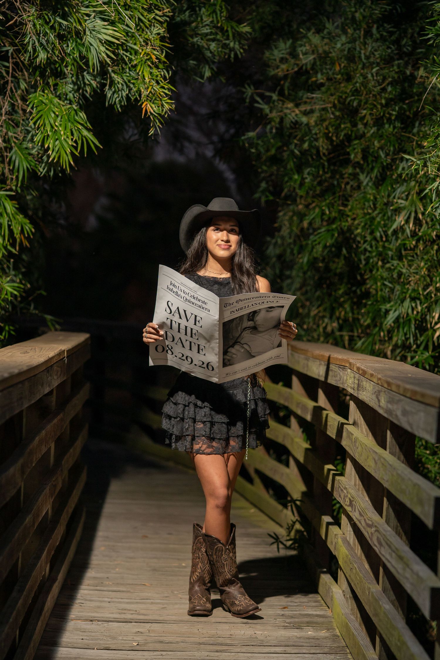 Woman in cowboy hat and boots holds Save the Date sign on wooden bridge surrounded by lush greenery.