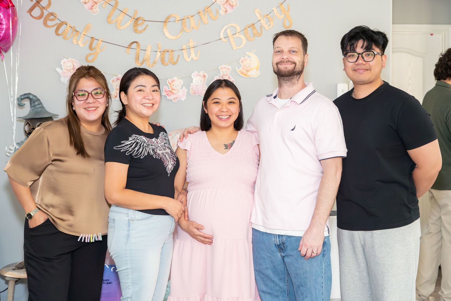 Group of five people posing at a baby shower, with a pregnant woman in pink at center.