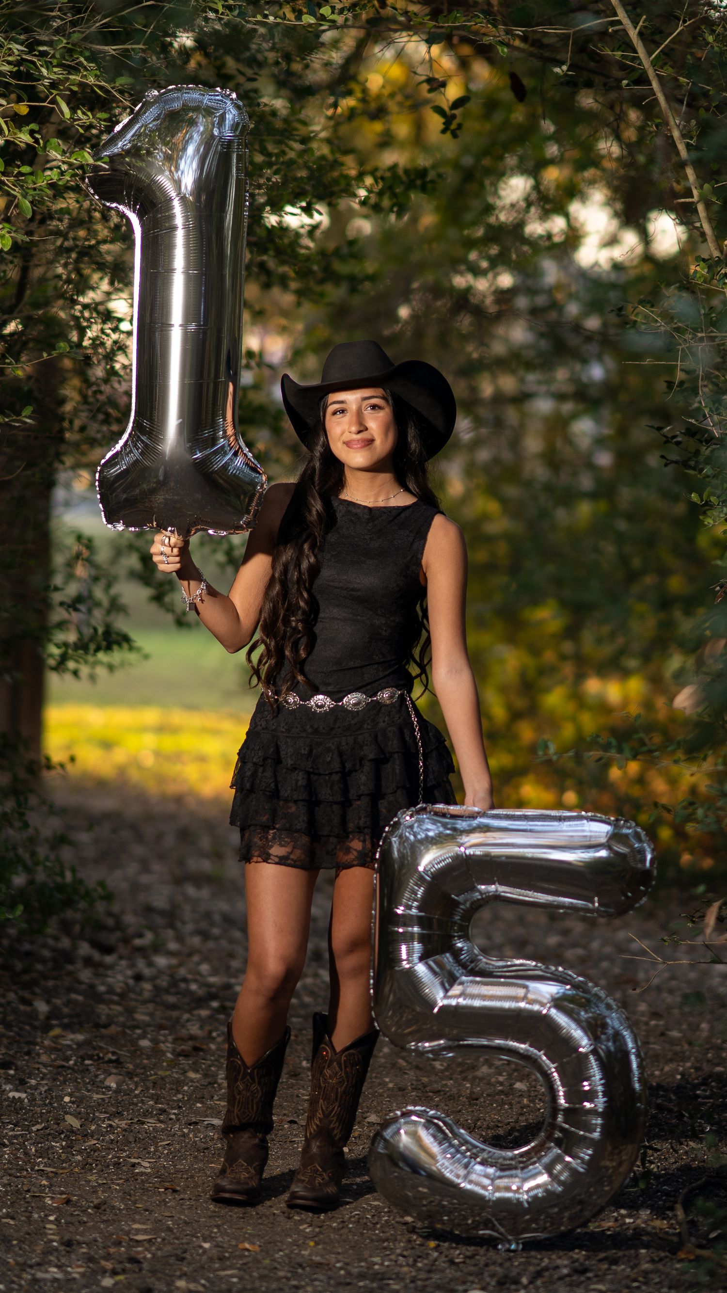 Teen girl in black cowgirl outfit holding silver number 15 balloons outdoors for quinceañera celebration.