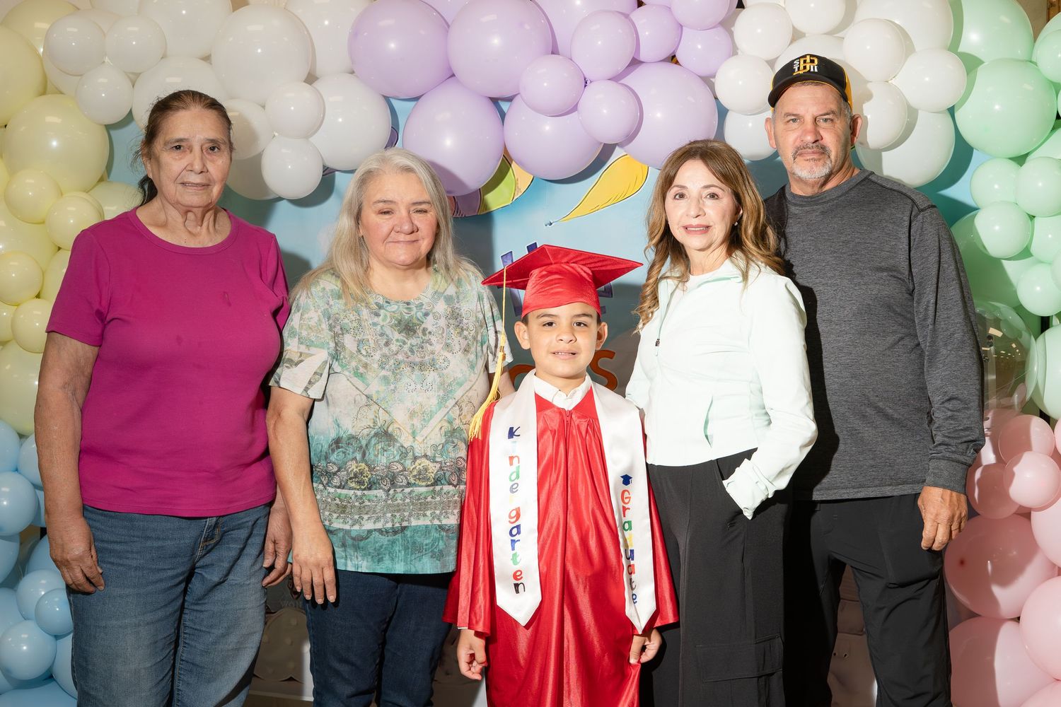 Young graduate in red cap and gown poses with four family members in front of colorful balloon backdrop.