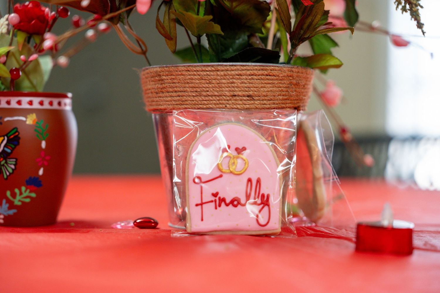 Pink decorated sugar cookie reading Finally in a clear bag beside a floral arrangement on a red table.