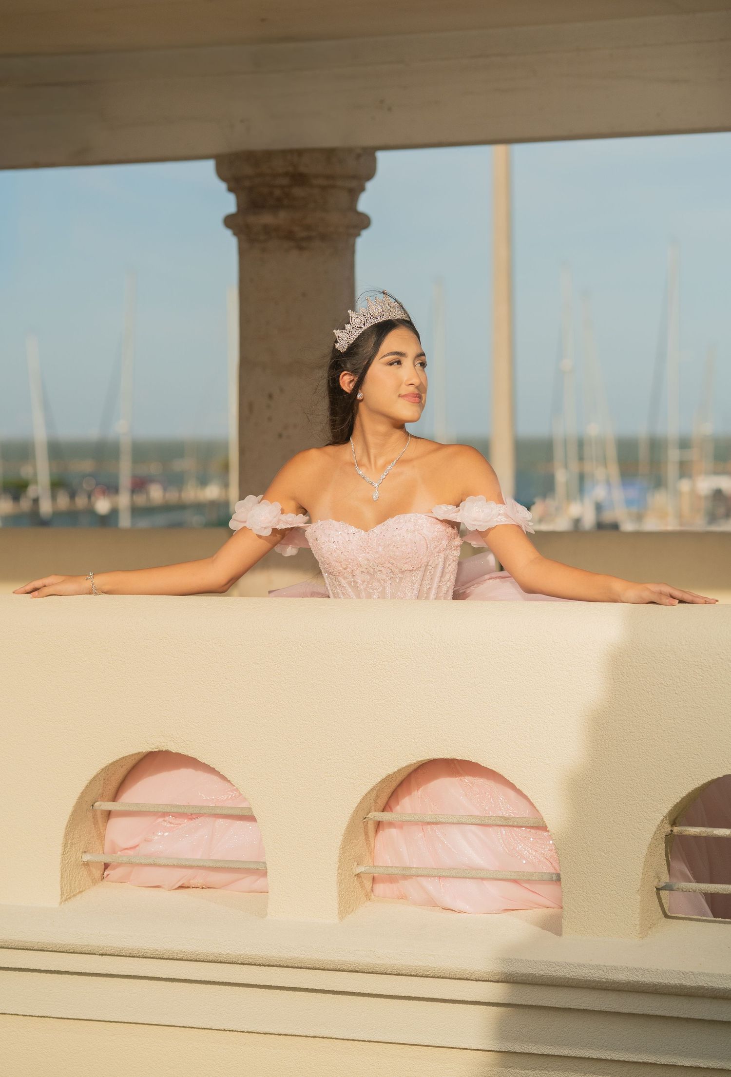 Elegant quinceañera in pink corset gown and tiara leans on balcony railing with marina backdrop.
