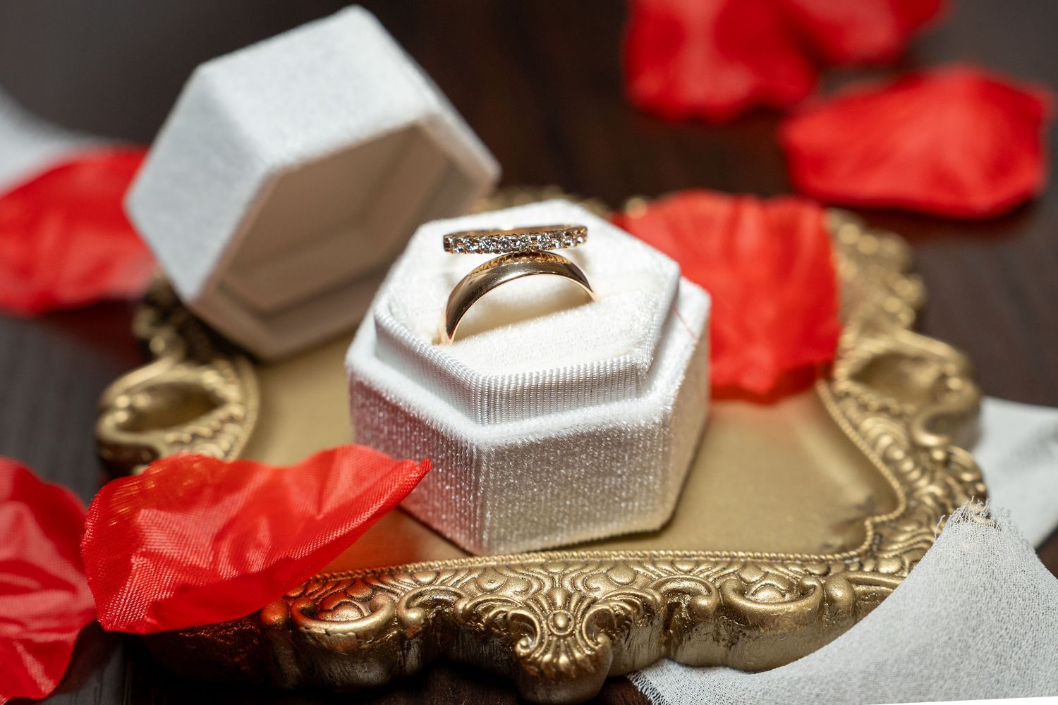Gold diamond ring displayed in white hexagonal jewelry box on ornate gold tray with red rose petals.