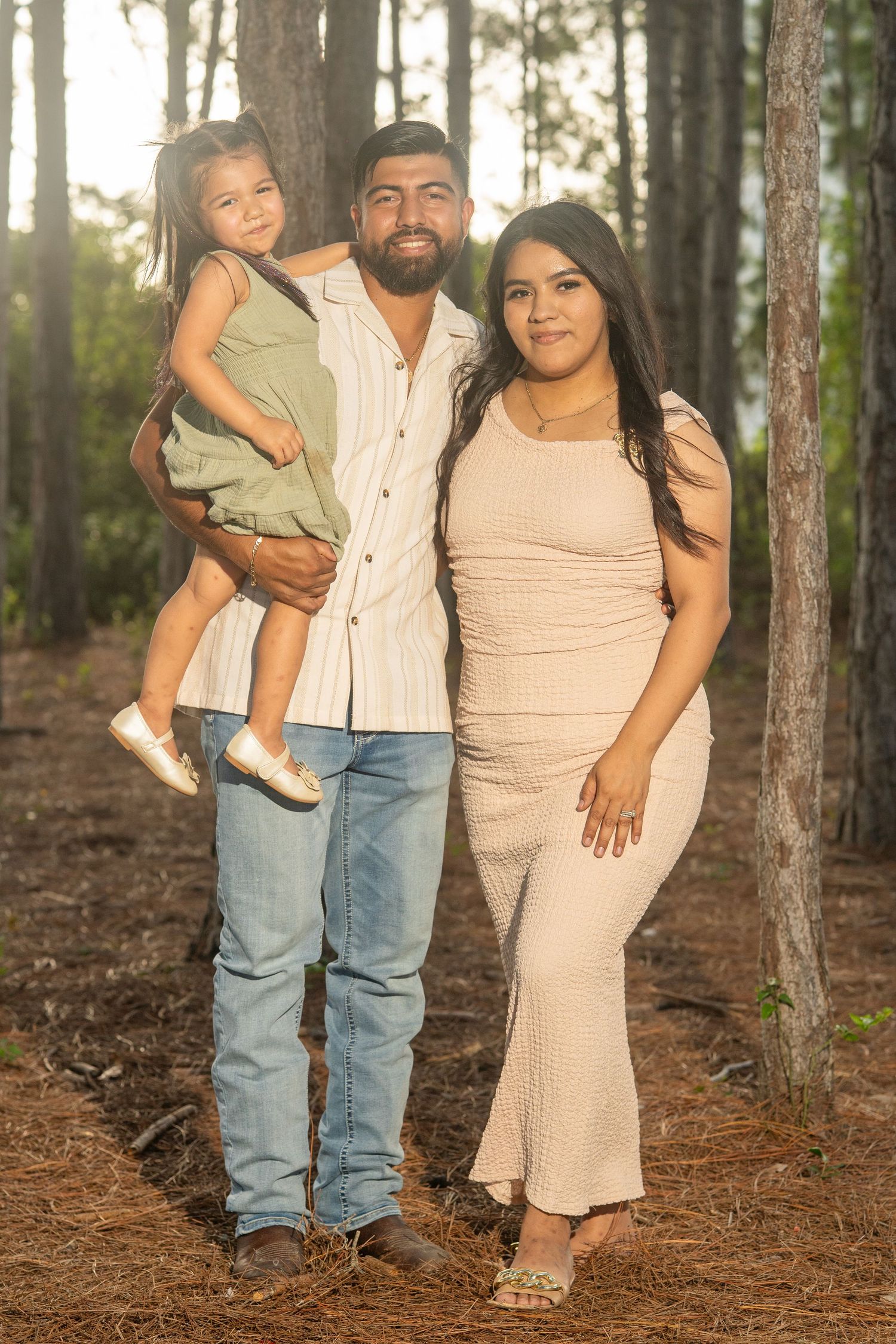 A family of three poses in a pine forest, with a man holding a toddler girl and a woman in a beige dress beside them.
