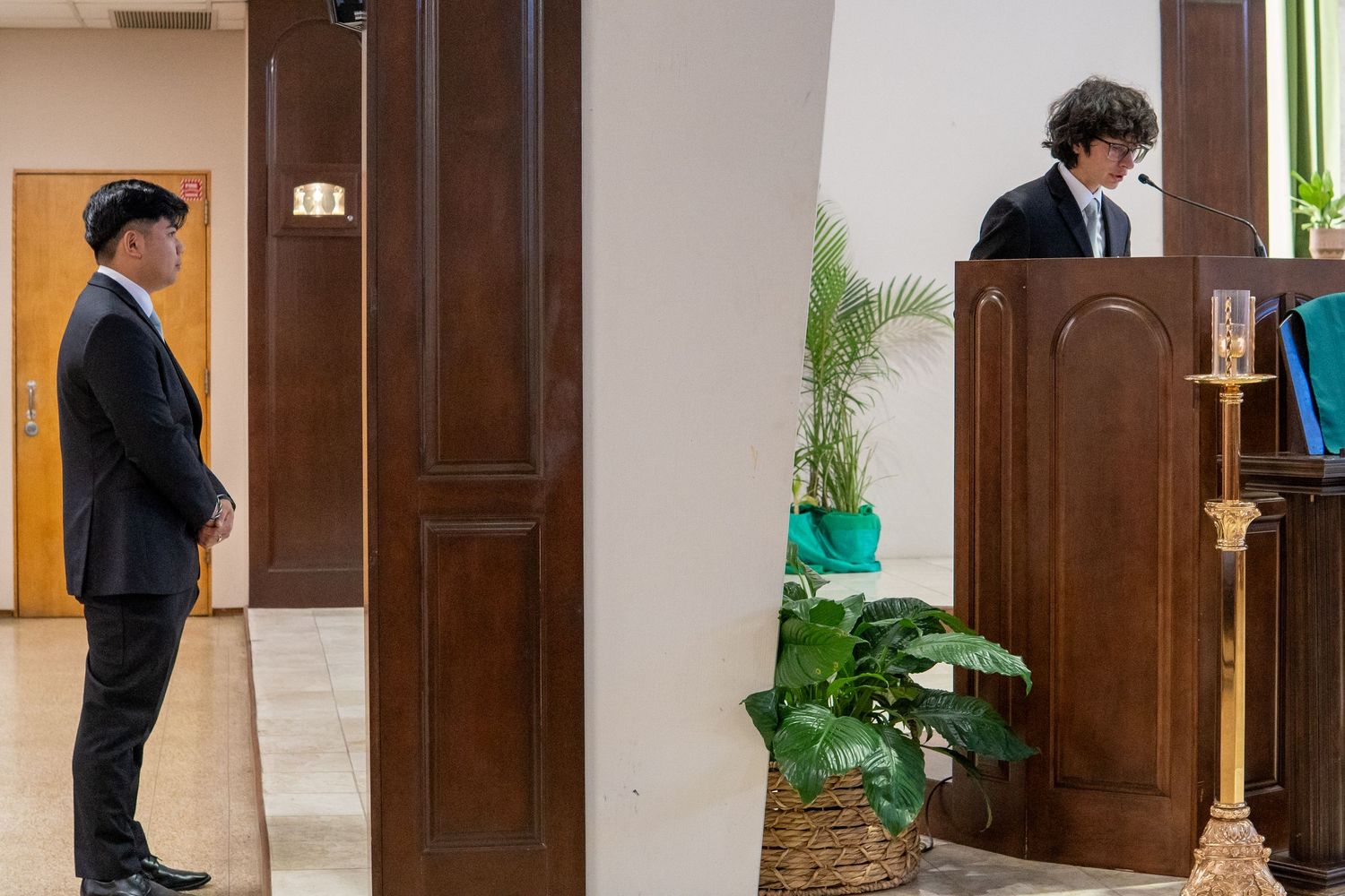 Two men in suits stand in a formal room, one at a wooden podium with plants and decor nearby.
