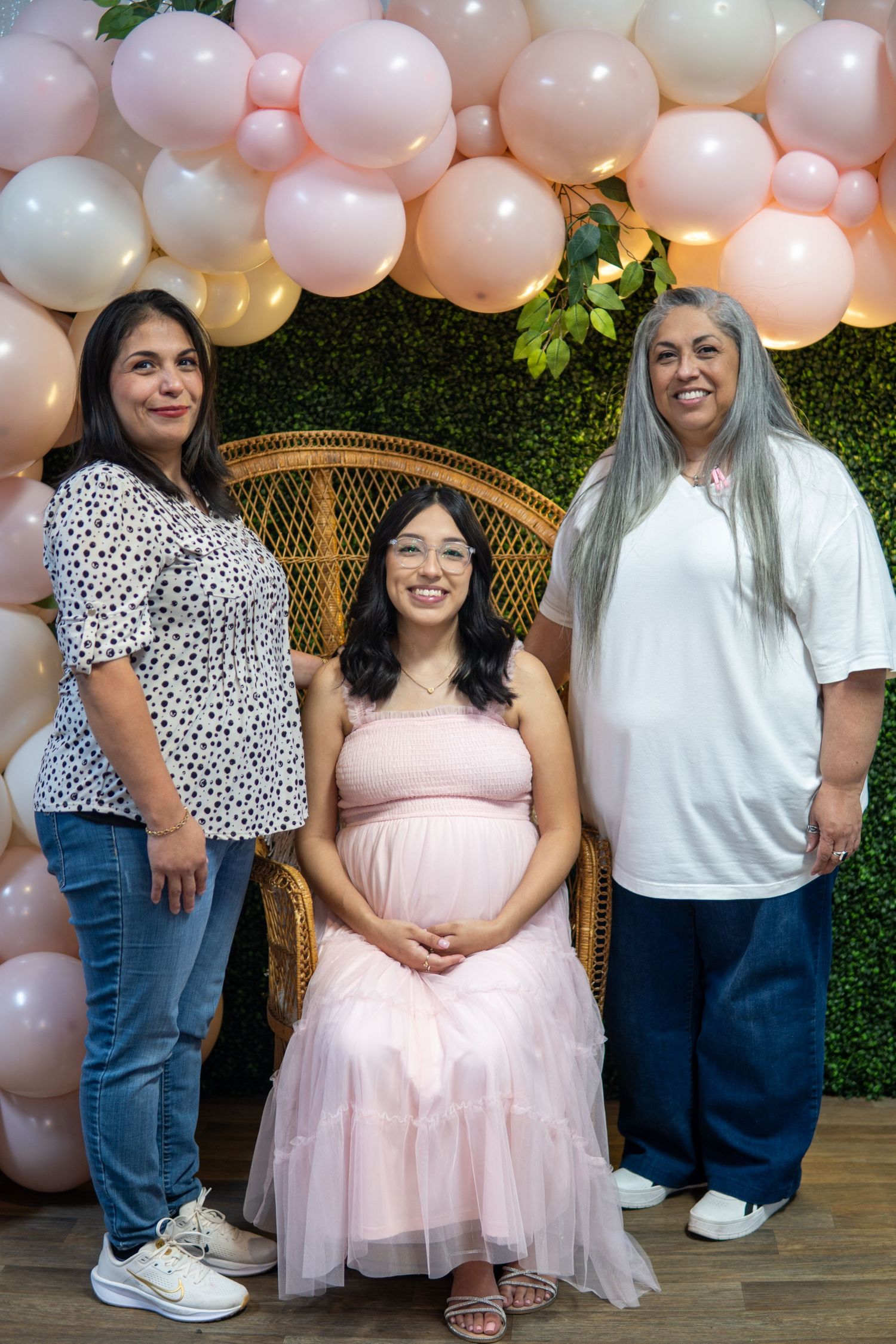 A baby shower celebration with pink balloons, a floral wall backdrop, and a wicker chair for photos at an indoor venue.