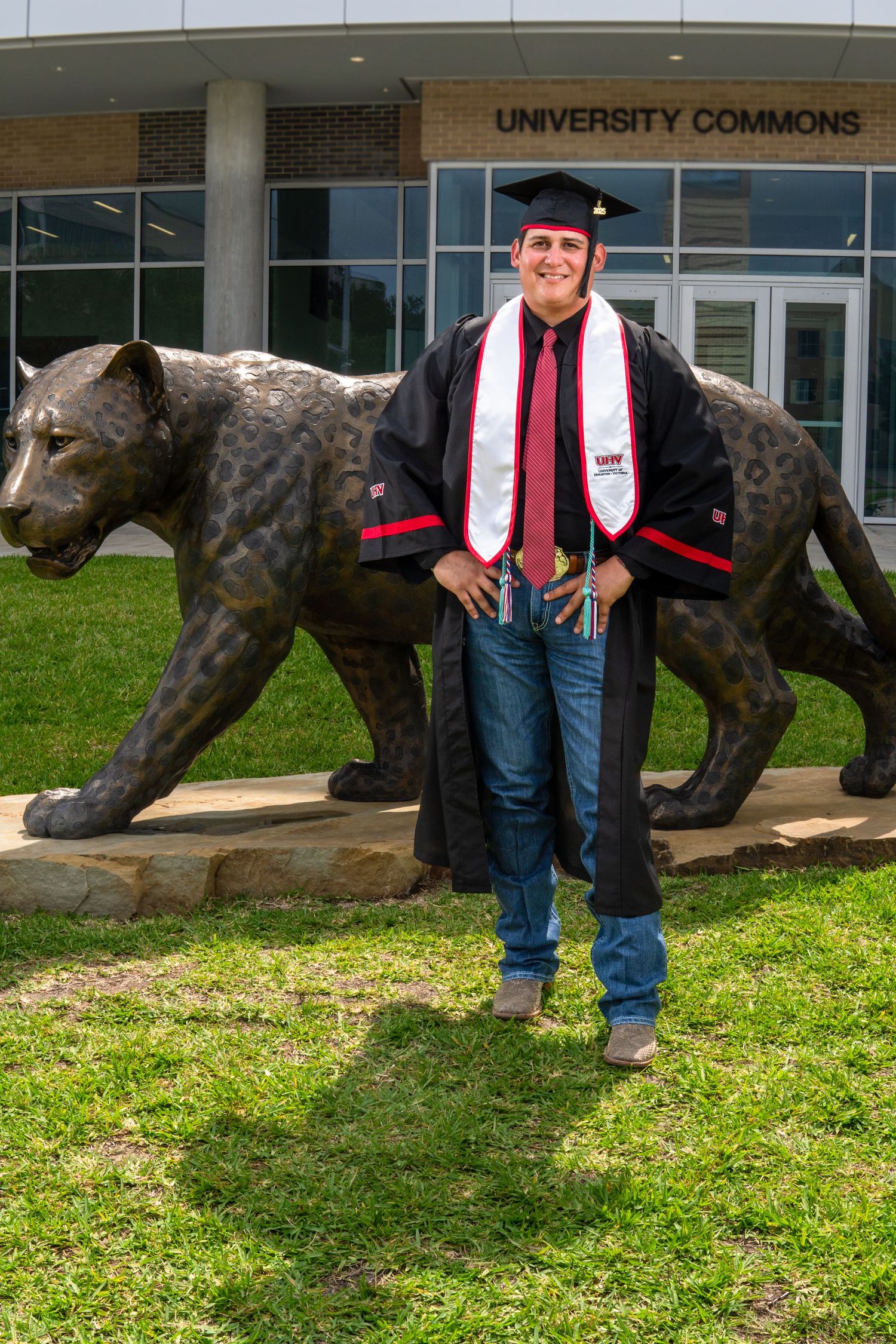Graduate in cap and gown poses next to wolf statue outside university commons building.