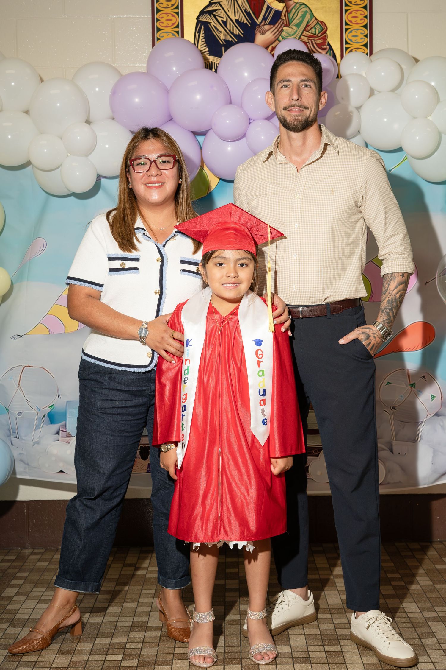A young girl in a red graduation cap and gown poses with two proud adults in front of a balloon backdrop.