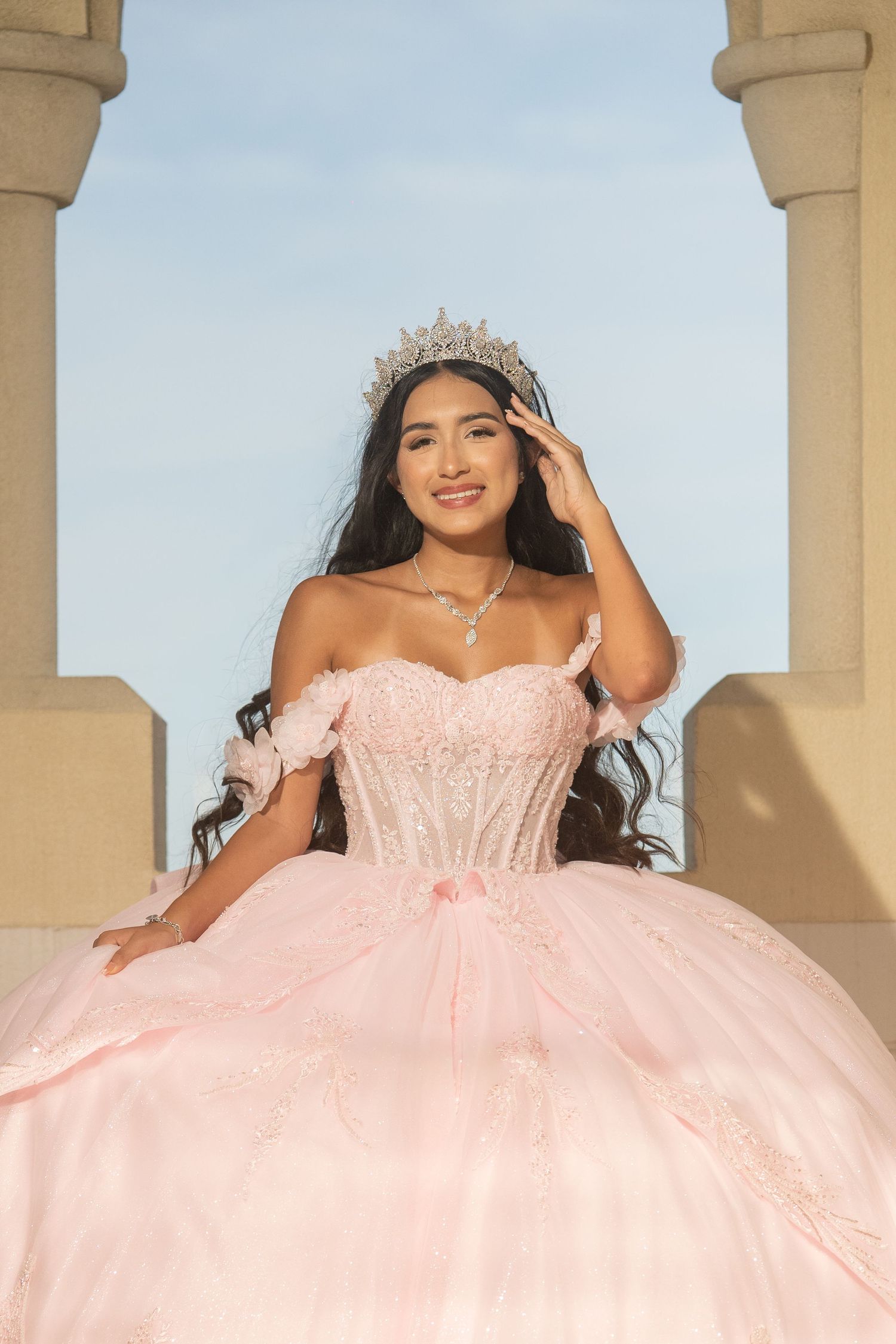 Smiling young woman in pink quinceañera gown and tiara poses between stone columns under blue sky.