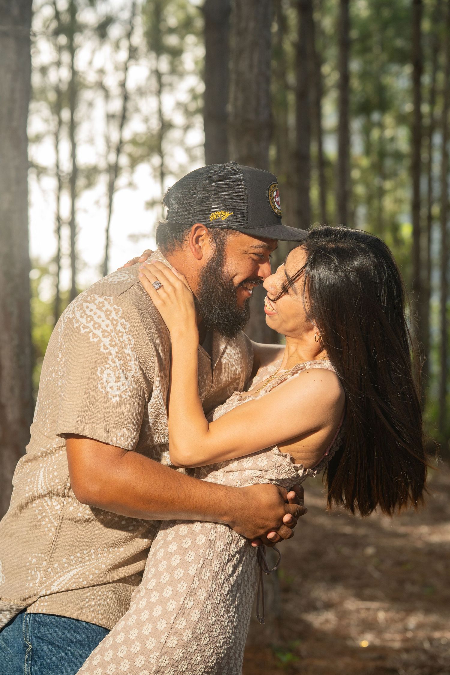 A couple shares a romantic embrace in a sunlit pine forest, smiling at each other in matching beige outfits.