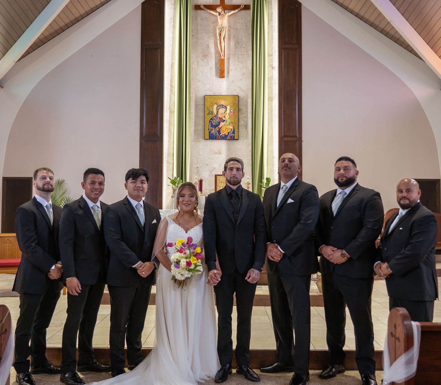 Wedding party poses in church with groom and bride holding colorful bouquet, flanked by six groomsmen in dark suits.