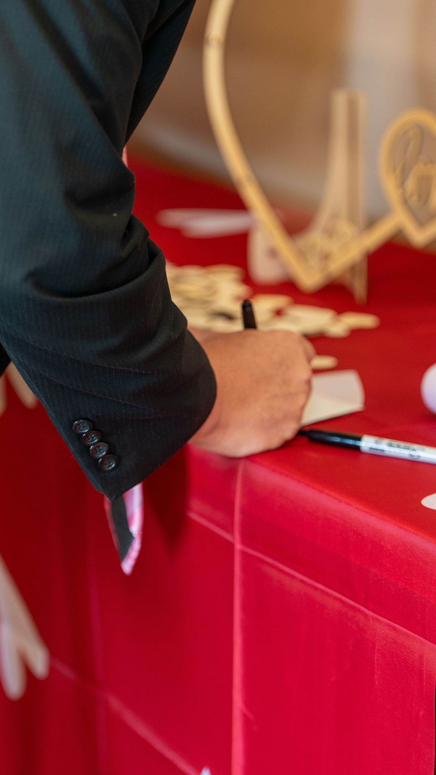 Guest in black suit signing wedding guestbook at red-draped table with wooden heart decorations.