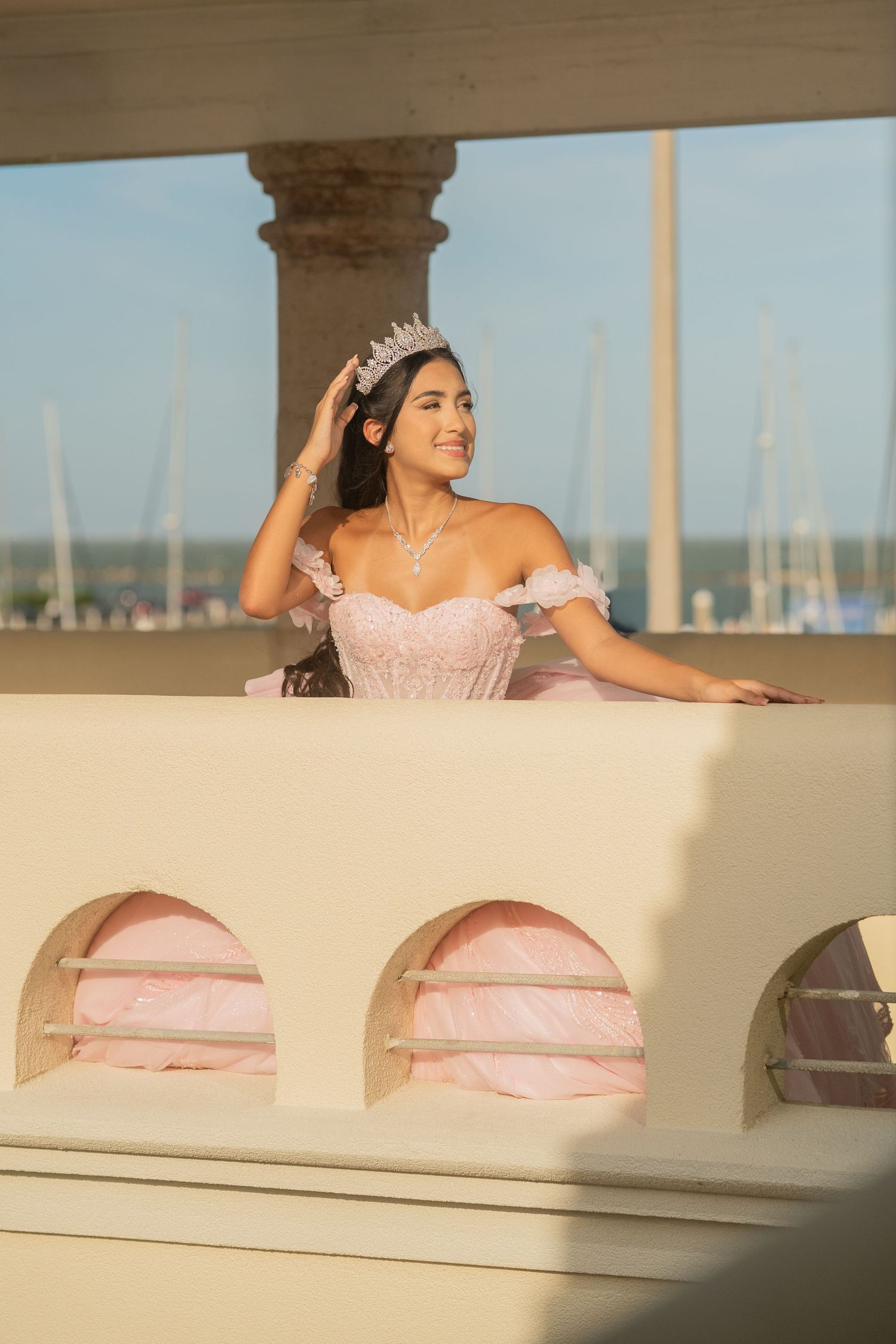 Young woman in pink quinceañera gown and tiara smiles on sunlit balcony overlooking a marina.