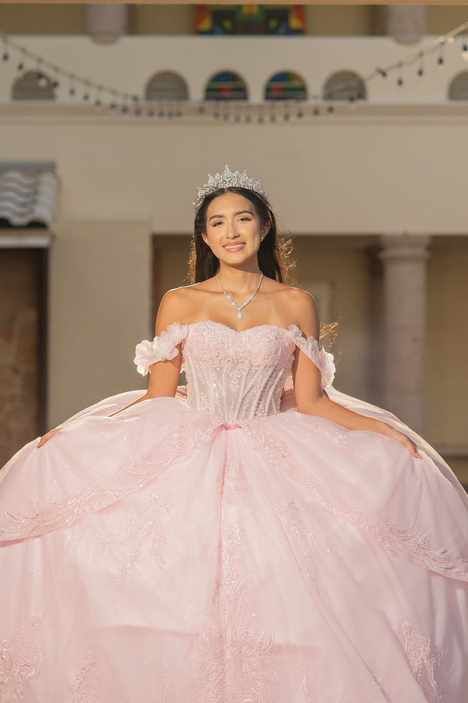 Beautiful young woman in a pink quinceañera ball gown with silver tiara smiling in a decorated venue.