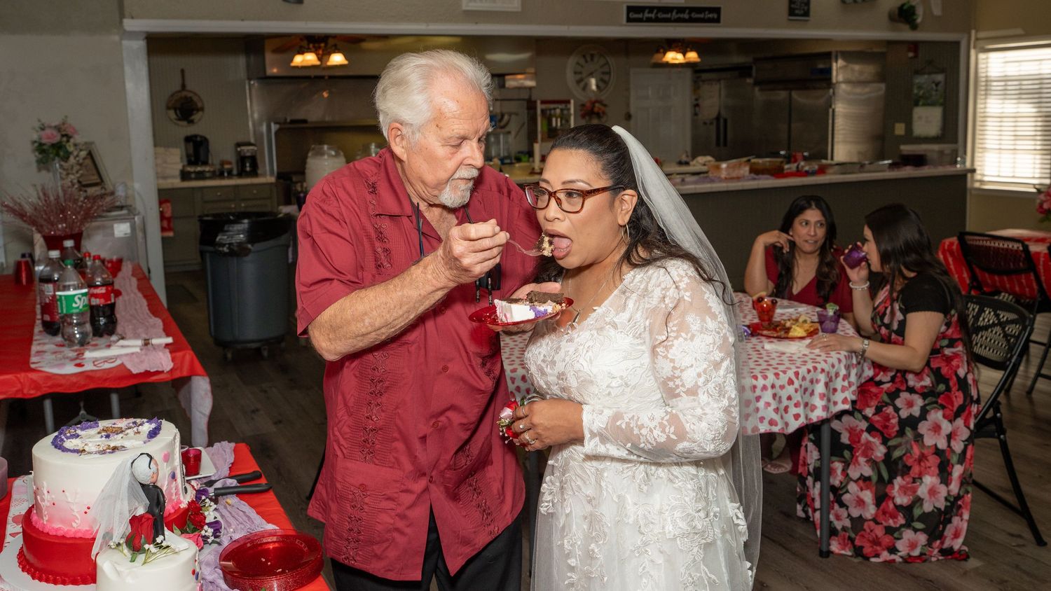 Elderly couple sharing wedding cake at reception, bride in white veil, groom in casual shirt, guests seated in background.