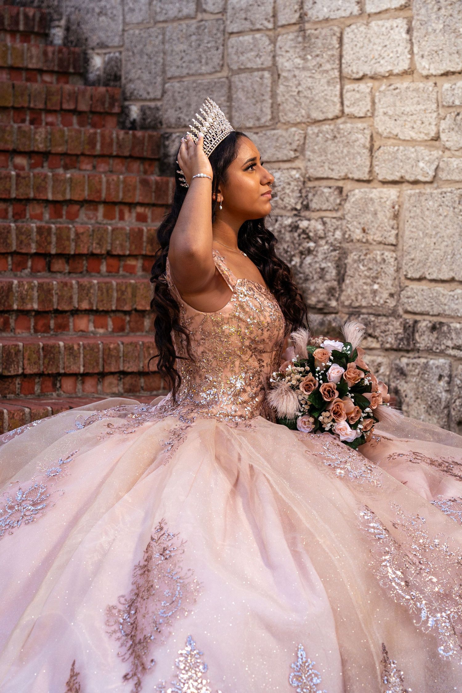 A stunning quinceañera gown in blush pink with intricate lace details and crystal embellishments against a stone wall backdrop.