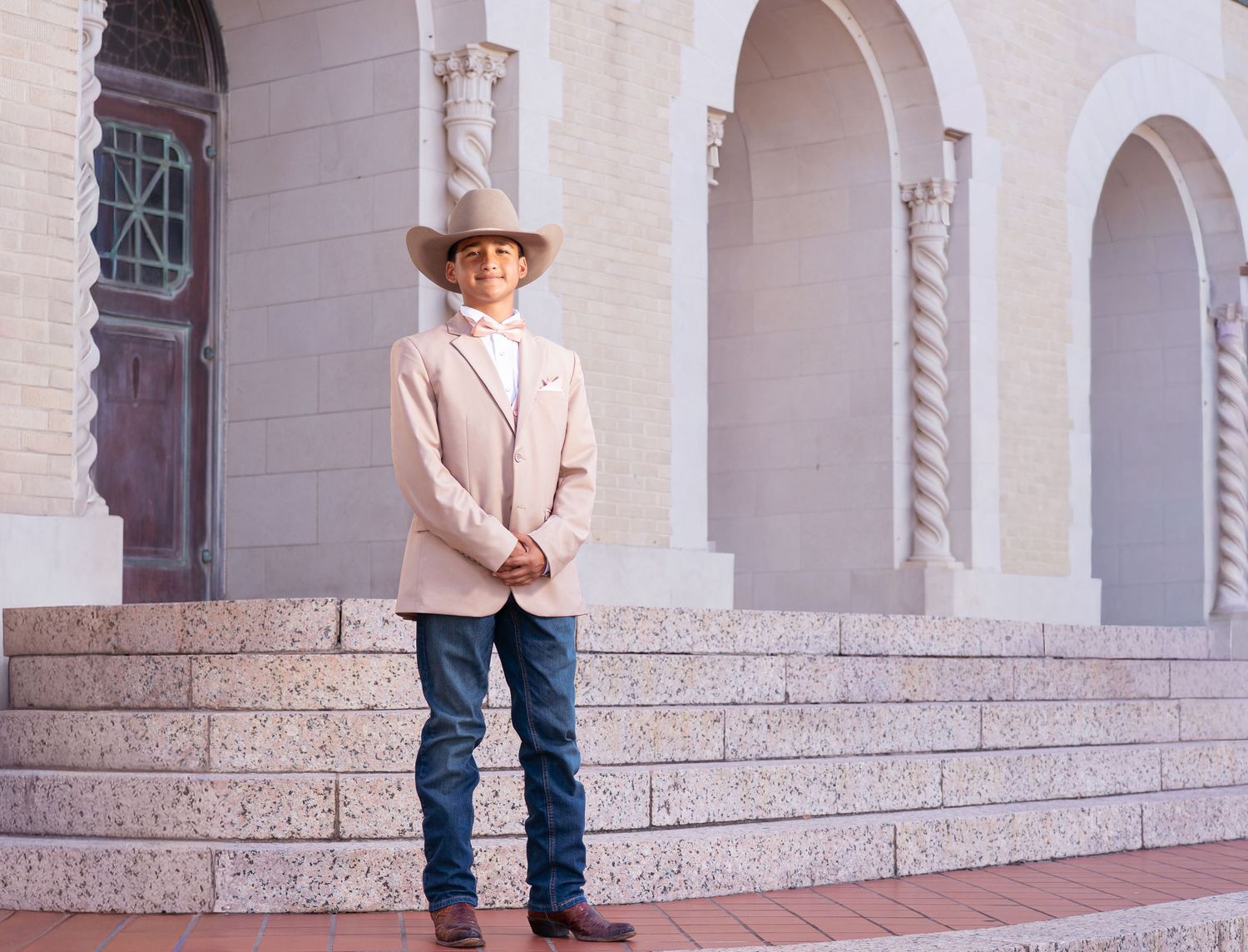 A person in a cowboy hat and light blazer stands on stone steps against a white arched building facade.