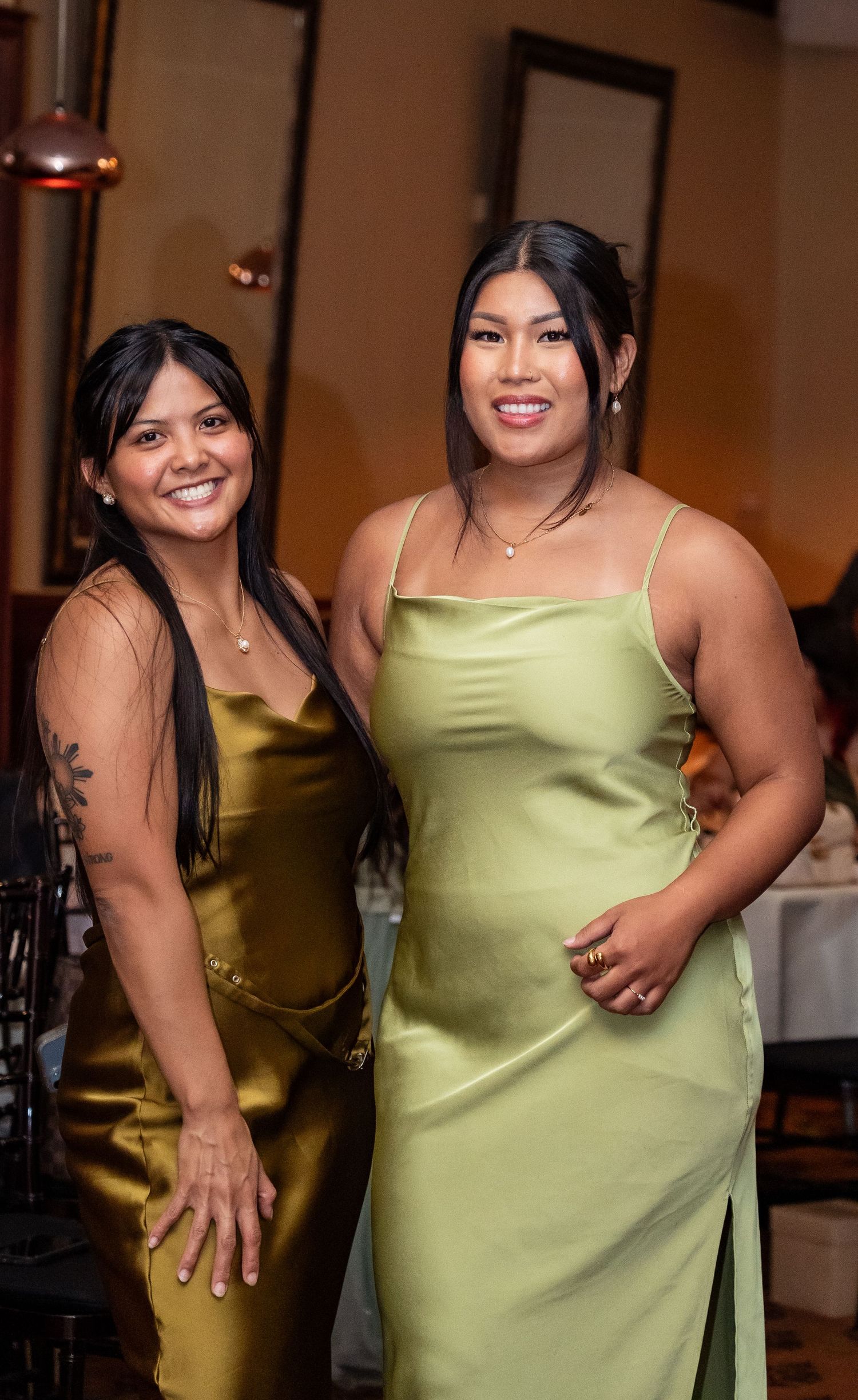 Two women pose together at a formal event wearing elegant satin slip dresses in gold and light green tones.