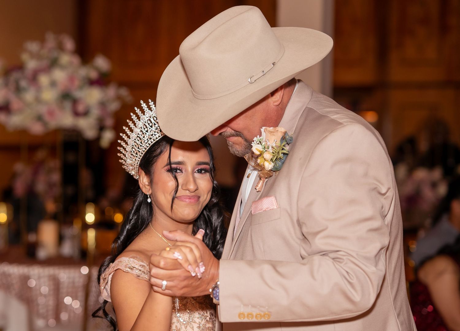 A young quinceañera in a tiara shares an emotional dance with a man in a tan cowboy hat and suit at her celebration.