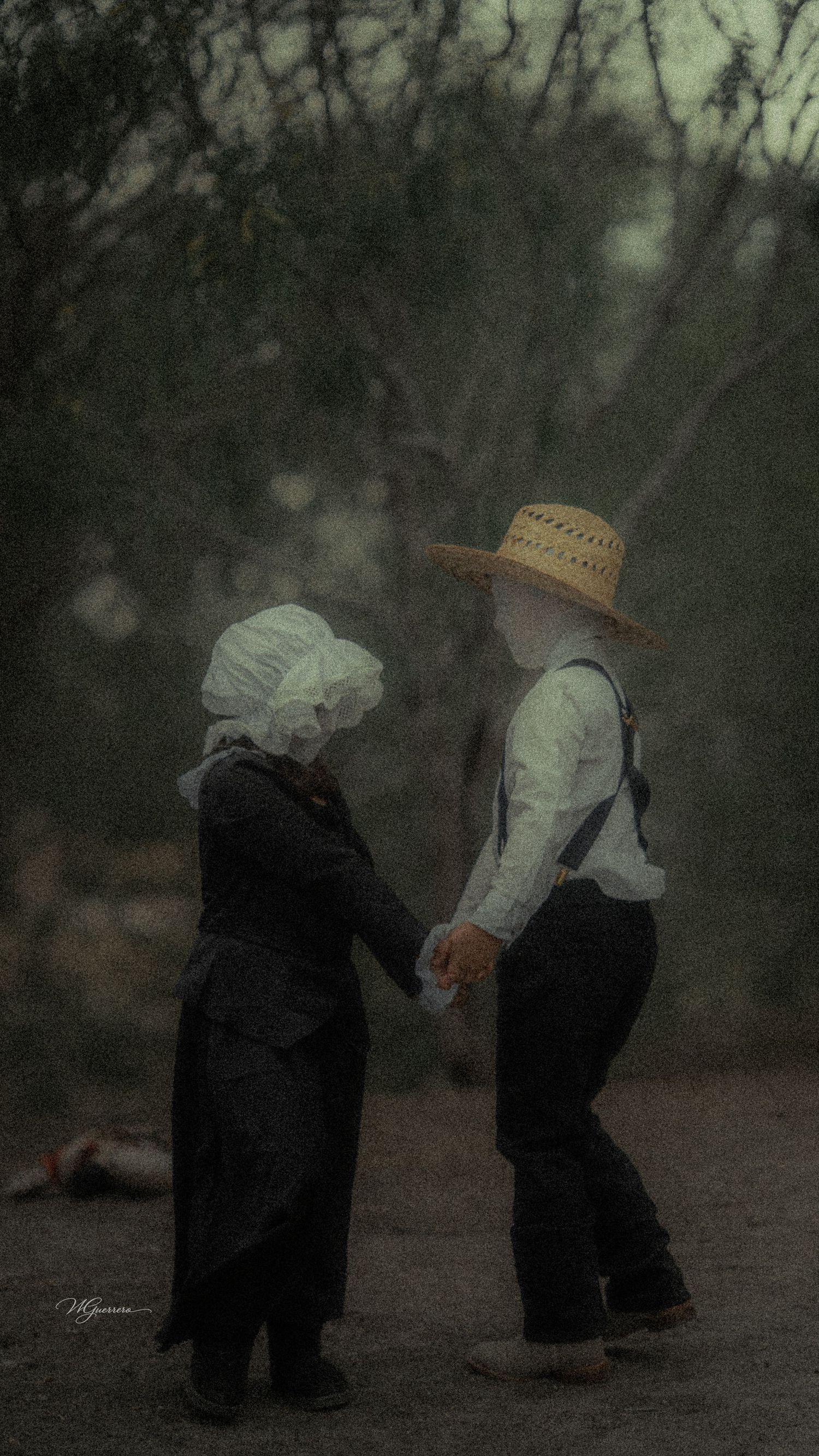 Two young children in vintage Amish-style clothing hold hands outdoors in a dark, misty setting.